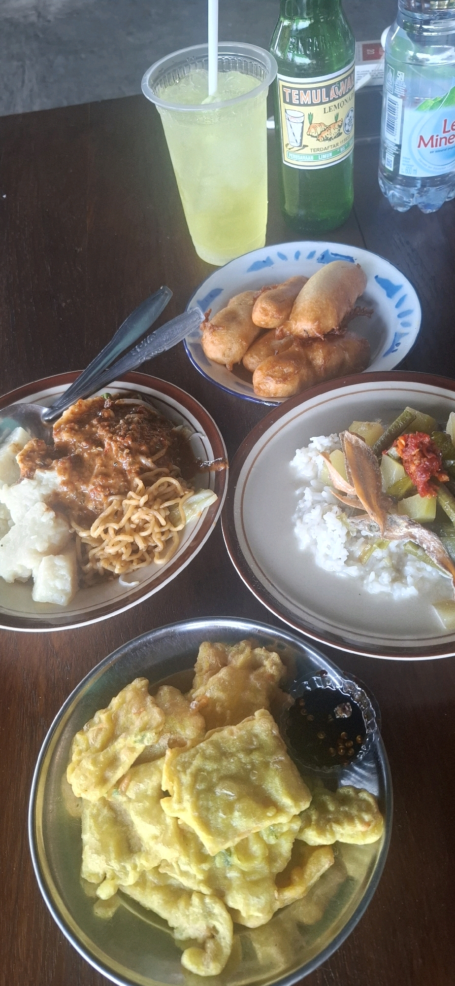 The image displays a close-up, eye-level view of a wooden table laden with food and drinks, likely taken at a food stall or eatery in Desa Sobokerto, Indonesia. In the foreground, a round metal plate is filled with several pieces of golden-brown, deep-fried tempeh, served with a small dish of dark dipping sauce. To the right, a white ceramic plate holds a serving of white rice topped with a stew containing green beans, potatoes, and what appears to be dried fish, garnished with red chili. To the left of the rice plate, another white plate contains a portion of rice accompanied by noodles and a rich, reddish-brown sauce with some vegetables. Behind these plates, a clear plastic cup holds a pale yellow, iced beverage with a straw. Next to it, a green glass bottle labeled "TEMULAWAK LEMONADE" is visible. Further to the right, a clear plastic bottle of water is present. The background is blurred, suggesting a shallow depth of field, and shows a dark, textured surface, possibly a wall or a dark wooden structure. The overall scene suggests a casual meal setting.