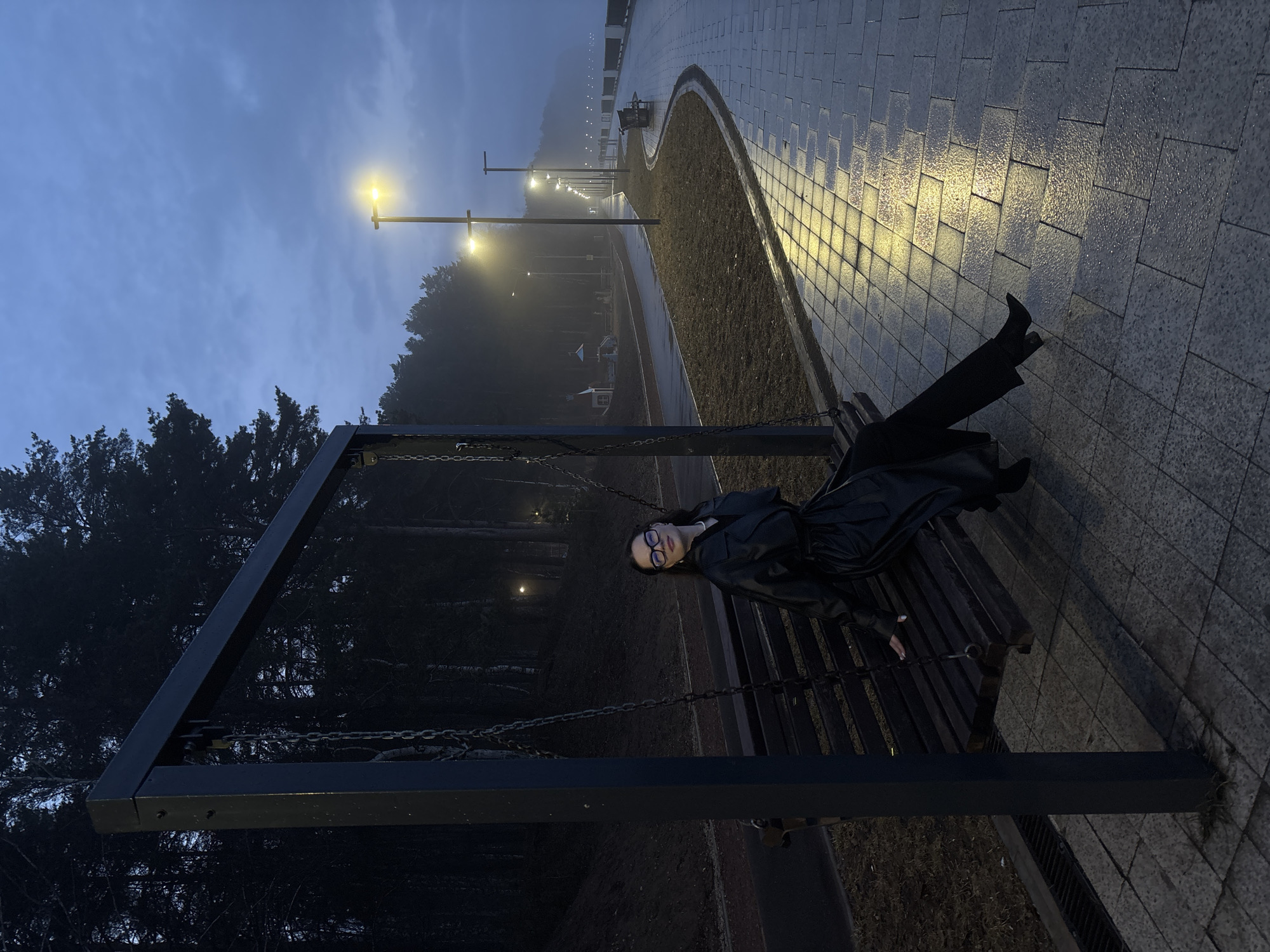 A medium-angle shot captures a woman reclining on a swing bench in Chaikovsky, Russia. The setting is a park or urban green space under a dusk or night sky, illuminated by evenly spaced streetlights lining a wet stone pathway. The woman is dressed in dark clothing, including what appears to be a trench coat and trousers, and wears glasses. She is positioned diagonally across the bench, facing towards the viewer. The bench is part of a swing structure with metal supports and chains. The pathway, made of rectangular stones, reflects the streetlight, indicating wet conditions. In the background, there are trees and indistinct structures obscured by mist or fog. The overall atmosphere is subdued and somewhat melancholic.