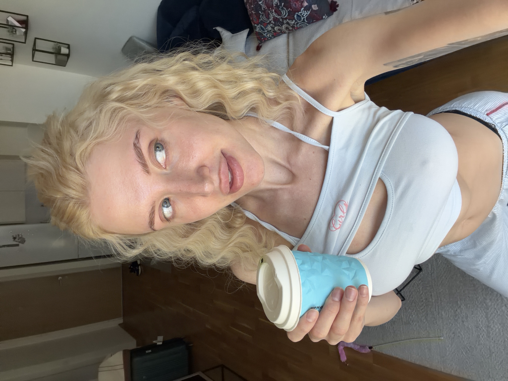 A woman with blonde curly hair and light eyes is holding a blue coffee cup in her right hand. She is wearing a white tank top with the word "Girls" printed on it, and striped shorts. She appears to be indoors, possibly in an apartment. In the background, there is a bed with pillows, a suitcase, and some furniture. It is difficult to determine the specific location in Belgrade, Serbia without more details.