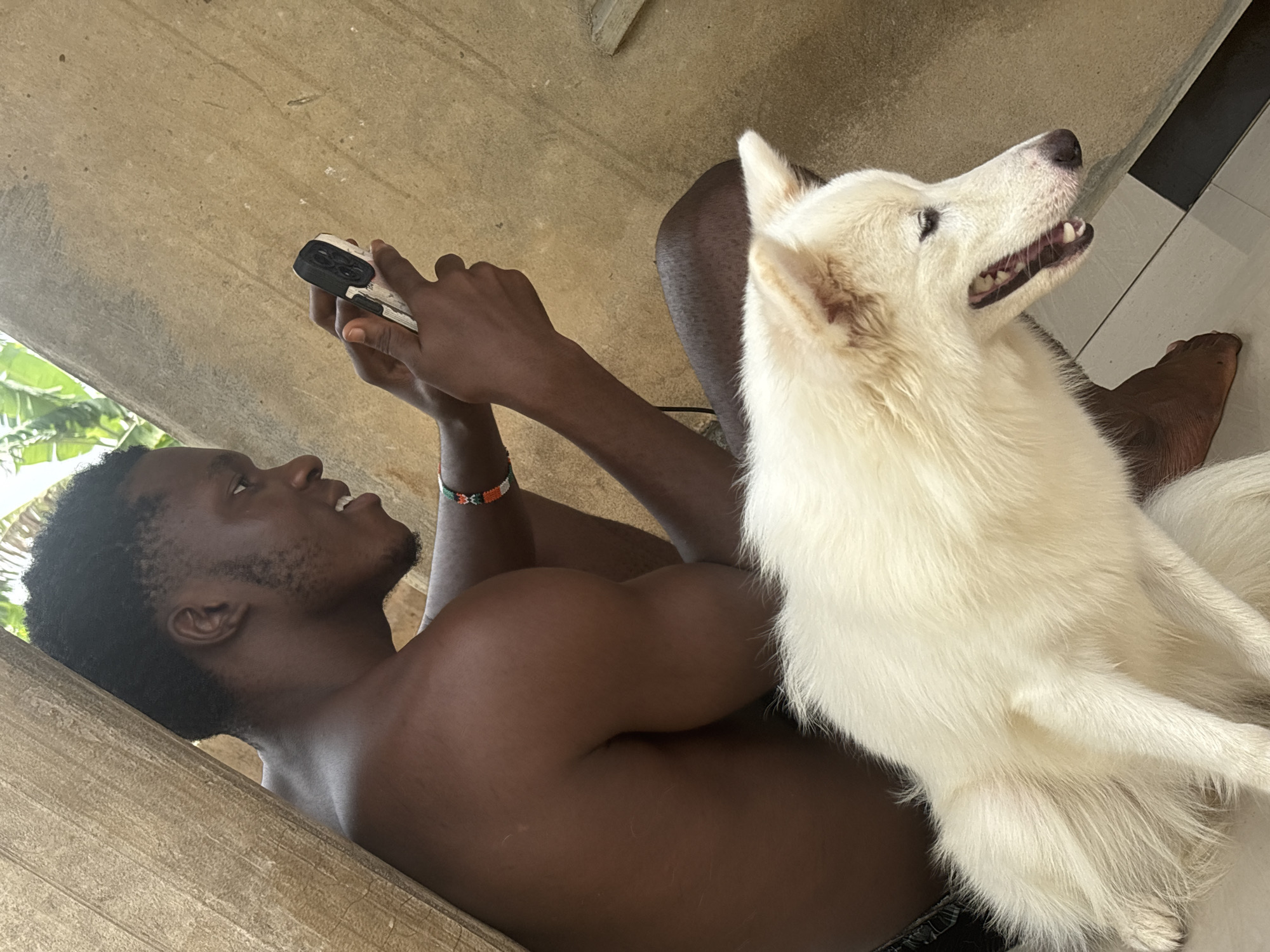 A man with dark skin lies on his back, propped against a weathered, light-colored wall or ledge. He's shirtless, with visible arm muscles, and is holding a smartphone above him, appearing to be taking a picture or video. He is wearing a beaded bracelet on his left wrist. His facial hair is trimmed close. Next to him, a large, white dog with thick fur is sitting upright and looking towards the right. The background shows glimpses of green foliage outside and a section of tiled flooring with black and white stripes in the upper right corner. The location and city are not discernible from the visual details in the image.