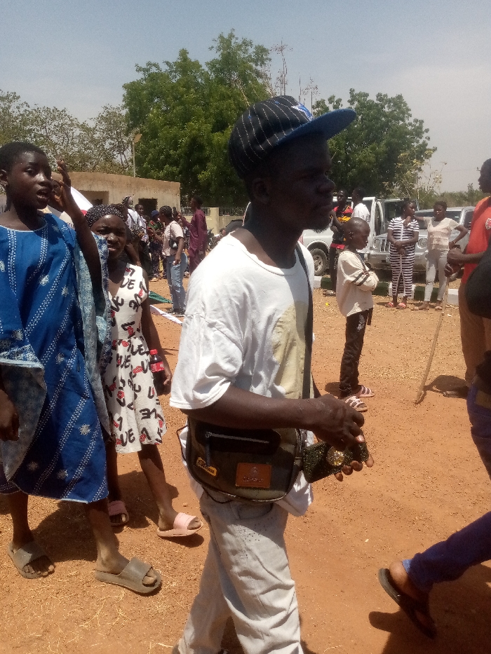 The image depicts an outdoor gathering in Kagoma, Nigeria, under bright daylight. The ground is reddish-brown dirt. In the foreground, from left to right, are three individuals. On the far left, a young person wearing a blue patterned tunic and tan slide sandals has their mouth open, appearing to speak, with their right hand raised. Next to them is a young girl in a white dress patterned with "LOVE" in black and red, holding a red Coca-Cola bottle in her left hand and wearing pink slide sandals. The central figure is an adult male, seen in profile, walking towards the left. He wears a white short-sleeved t-shirt, light-colored pants, and a black and blue striped baseball cap worn backward. A dark green fanny pack is strapped across his waist. He holds a dark, rectangular object, consistent with a mobile phone, in his right hand.
In the mid-ground and background, numerous other individuals are gathered. A light-brown, single-story building is visible to the far left. Several white utility vehicles or SUVs are parked in the mid-ground to the right, with people standing near them, including a child in a light-colored top and dark pants. One individual further right holds a long wooden stick or staff. Green trees and shrubs are present in the background against a pale blue sky.