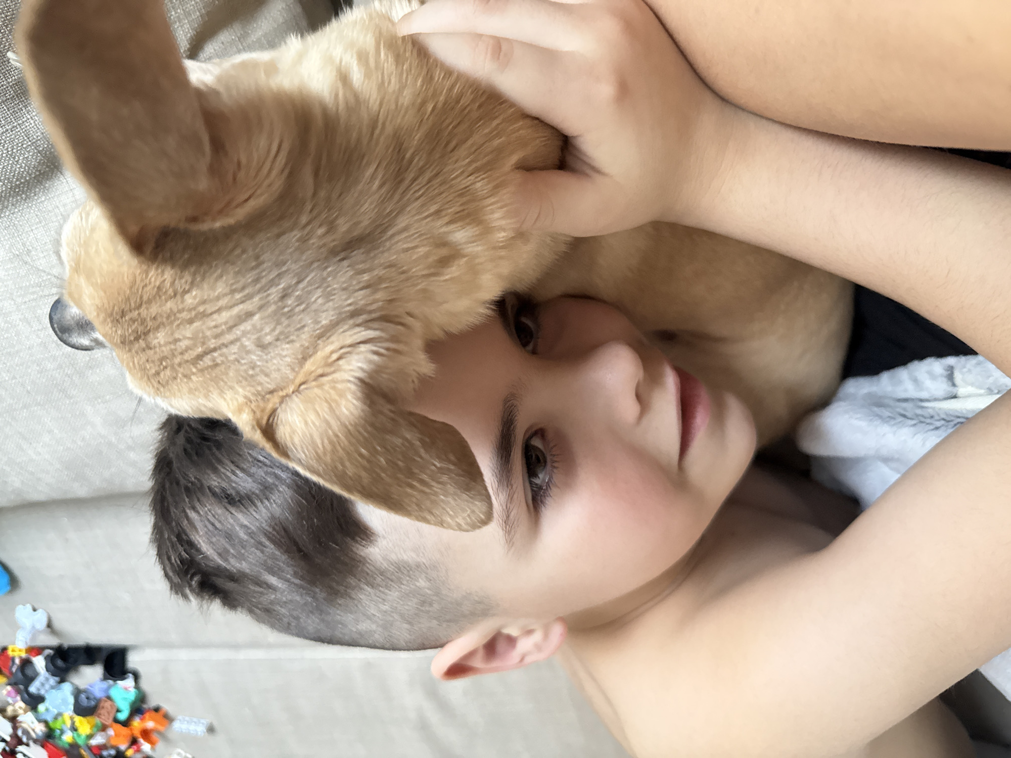 The image captures a tender moment between a child and a dog, viewed from an overhead perspective and rotated slightly to the side. A young child, appearing to be a boy, is lying down, looking up and directly towards the viewer with warm brown eyes and a soft, gentle expression. He has short, dark hair that is closely cropped on the sides. His bare shoulder and part of his chest are visible, suggesting he is shirtless.
Resting its head gently on the child's head and neck area is a light brown or tan short-haired dog. The dog's fur appears soft and clean. A human hand, likely the child's, is visible near the dog's head, appearing to be petting or holding the dog affectionately. The dog's ear is upright, and its snout is visible close to the child's face, indicating closeness and comfort between the two.
The subjects are positioned on what appears to be a light beige or off-white upholstered surface, possibly a couch or a bed, suggesting an indoor setting. In the bottom left corner of the image, a scattering of colorful small plastic toy pieces, possibly building blocks like Lego, can be seen on the floor next to the furniture. The lighting in the image is soft and natural, suggesting it is daytime indoors. The overall atmosphere conveyed is one of warmth, affection, and peaceful companionship.
