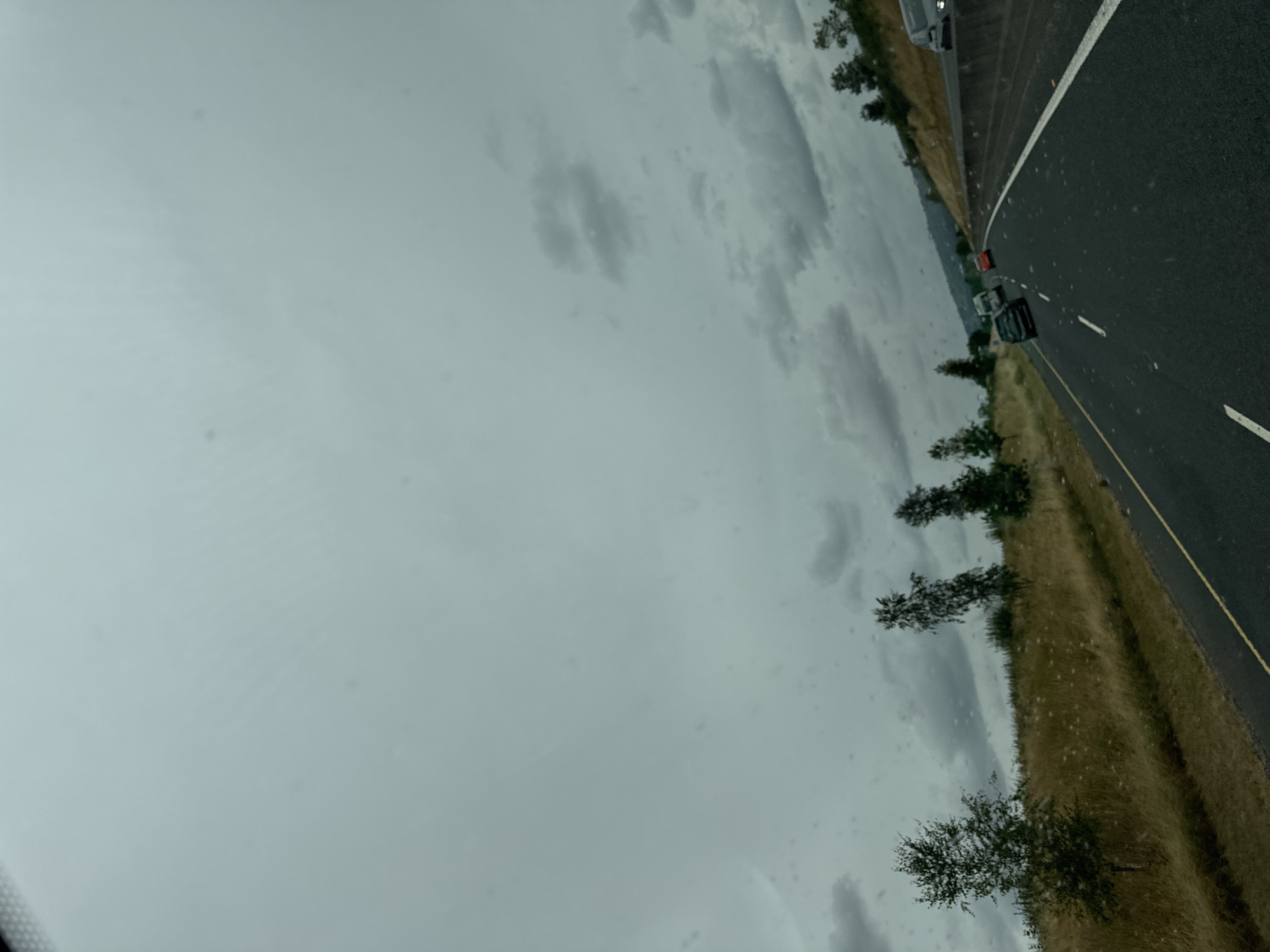 The image is a first-person perspective, likely from a vehicle moving along a highway. The sky is overcast with scattered clouds, suggesting a possibly dull or rainy day. On the left side of the road, there is a grassy embankment with several trees and dry grass. The road itself is asphalt, with a yellow line marking the edge and white dashed lines indicating lanes. Several cars are visible on the highway, with one car in the foreground and others further ahead. The overall environment suggests a rural or semi-rural setting. The location is identified as Tinryland, Ireland.