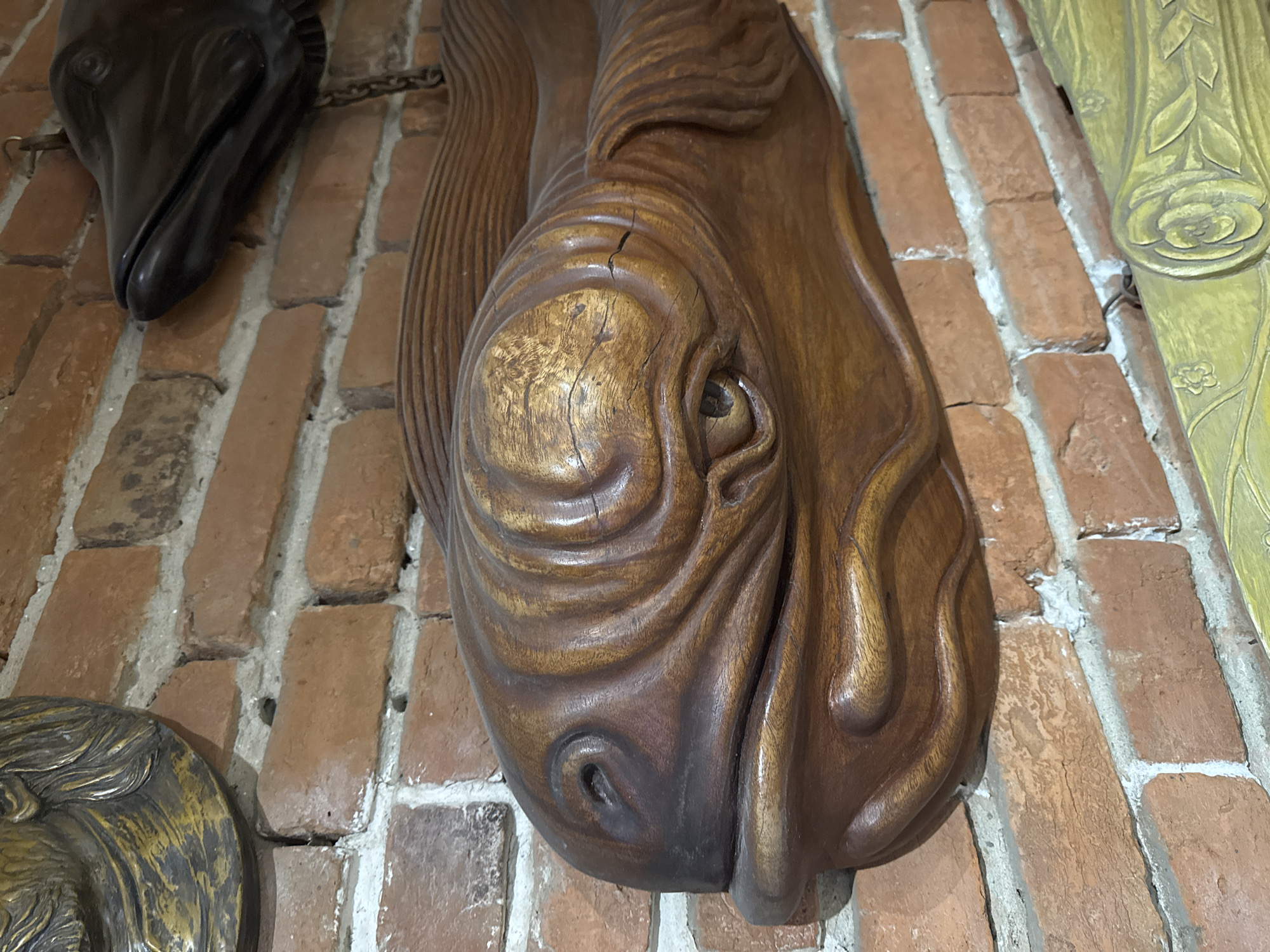 The image shows a close-up of a wooden fish sculpture mounted on a brick wall. The fish sculpture is brown and has visible grain and texture, with carved details suggesting scales and fins. Its eye is a dark circular recess, and its mouth is slightly open. To the upper left, a portion of a gold-colored, ornate decorative object is visible, possibly a mask or a relief. To the upper right, a dark, smooth sculpture of a fish head with an open mouth is also attached to the wall, with a chain visible. The brick wall is made of reddish-brown bricks with white mortar. A portion of a yellow, carved decorative element is visible at the bottom of the frame, featuring floral motifs. The overall impression is of decorative objects displayed indoors, possibly in a rustic or historically themed setting. No specific location in Kolomyia, Ukraine, can be identified from the image itself.