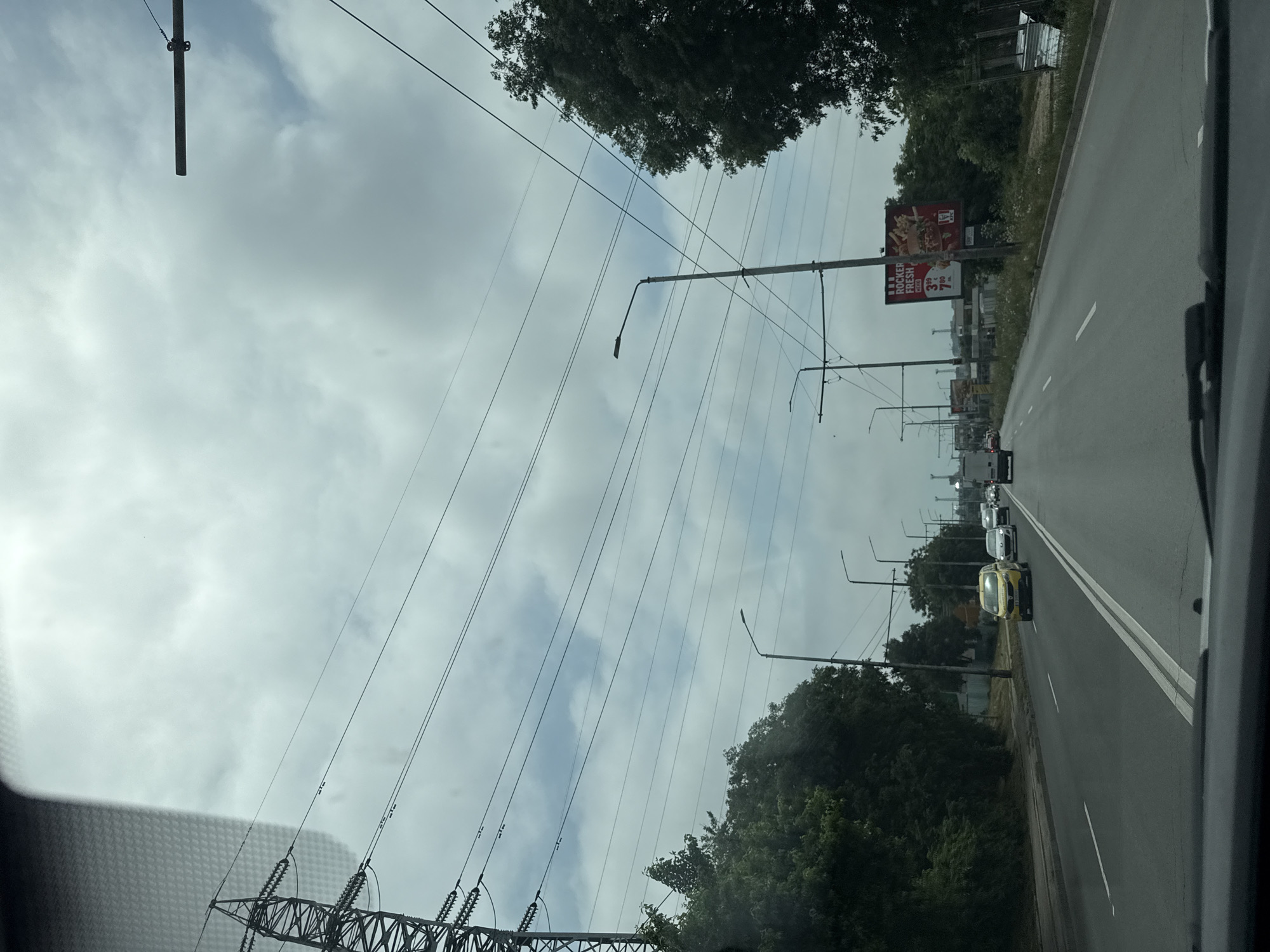The image captures a roadside scene from the perspective of a vehicle, looking ahead down a multi-lane asphalt road under an overcast sky. The sky is predominantly grey with visible cloud formations.
Numerous utility poles and streetlights line both sides of the road, supporting a complex network of overhead power lines that crisscross the frame. On the left side, a large, metal lattice-style power transmission tower is prominently visible, connected to some of these lines.
The road features clear white lane markings, including a double solid line separating opposing directions and single solid and dashed lines defining lanes. Several vehicles are present on the road, indicating active traffic. A distinctive yellow car is in the foreground, in the left lane, with a white truck and other cars further down the road.
To the right of the road, beyond a dirt verge, there are trees and bushes, and a large billboard advertising "ROCKER FRESH" with prices "3.99 BGN" and "7.00 BGN." A partially visible logo suggests it belongs to the fast-food chain KFC. More trees and greenery flank the road on the left side as well. The presence of "BGN" (Bulgarian Lev) on the billboard confirms the location as Varna, Bulgaria. The scene depicts a typical daytime urban or suburban road, likely on a cloudy day.