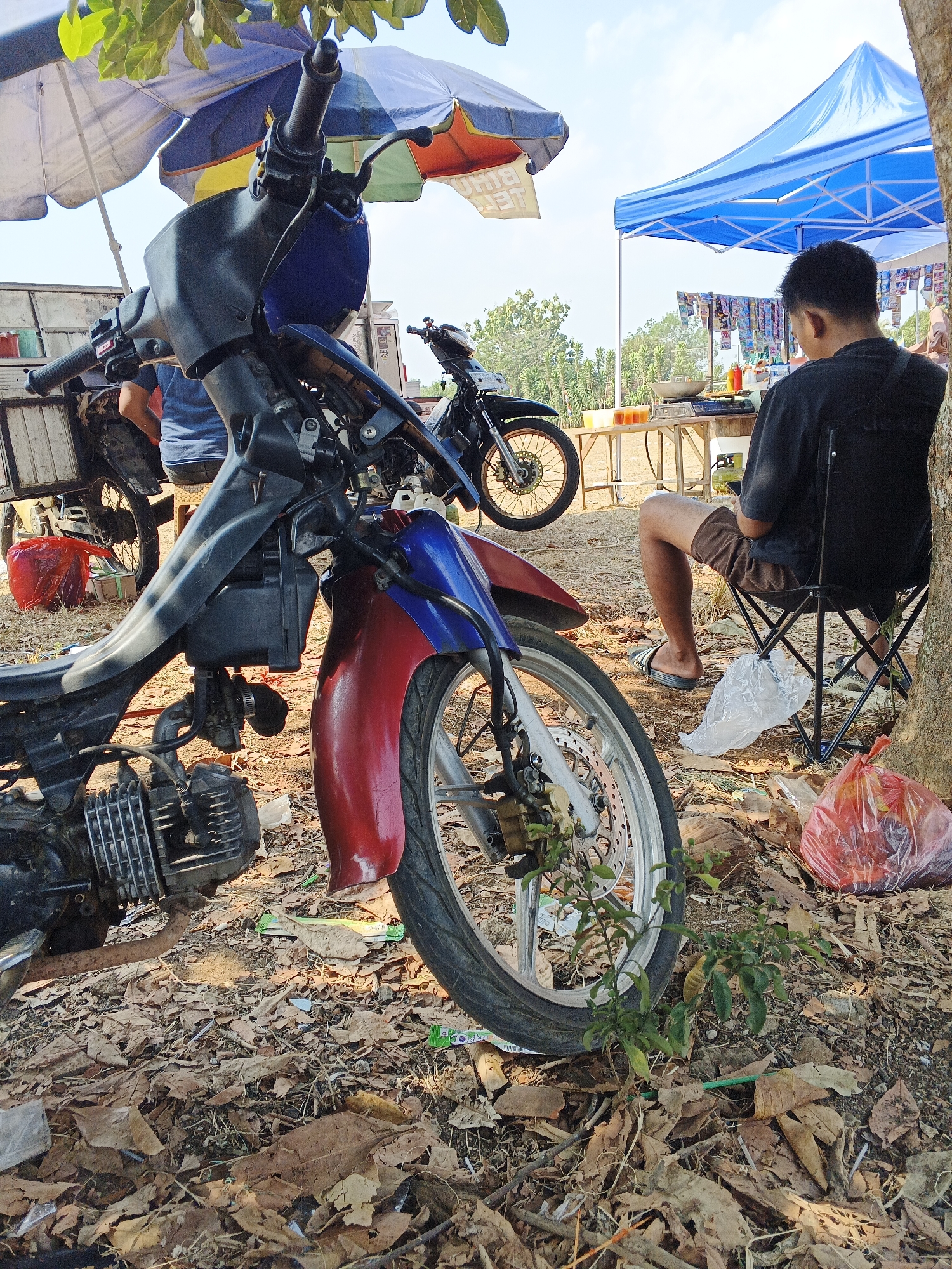An outdoor scene in Warungkiara, Indonesia depicts a small roadside market under a clear sky. The foreground is dominated by the front portion of a disassembled motorcycle, its red and blue fairing prominently displayed. Its engine, exhaust pipe, and front wheel are visible, resting on a ground covered in dry leaves and debris. To the left, another motorcycle is partially visible, parked behind an open-sided vehicle.
In the mid-ground, a blue canopy tent is set up, beneath which a person is seated in a folding chair, facing away from the camera. This individual wears a black t-shirt and brown shorts. In front of the tent, a wooden table displays a wok, some cups of drinks, and various packaged goods. A white plastic bag lies on the ground near the seated person, and a red plastic bag filled with items is on the ground to the right.
Further back, another canopy, this one a mix of blue, yellow, and red, provides shade for more stalls. In the background, lush green trees and foliage are visible, suggesting a rural or semi-rural setting. The overall atmosphere is one of casual commerce and everyday life in a tropical environment.