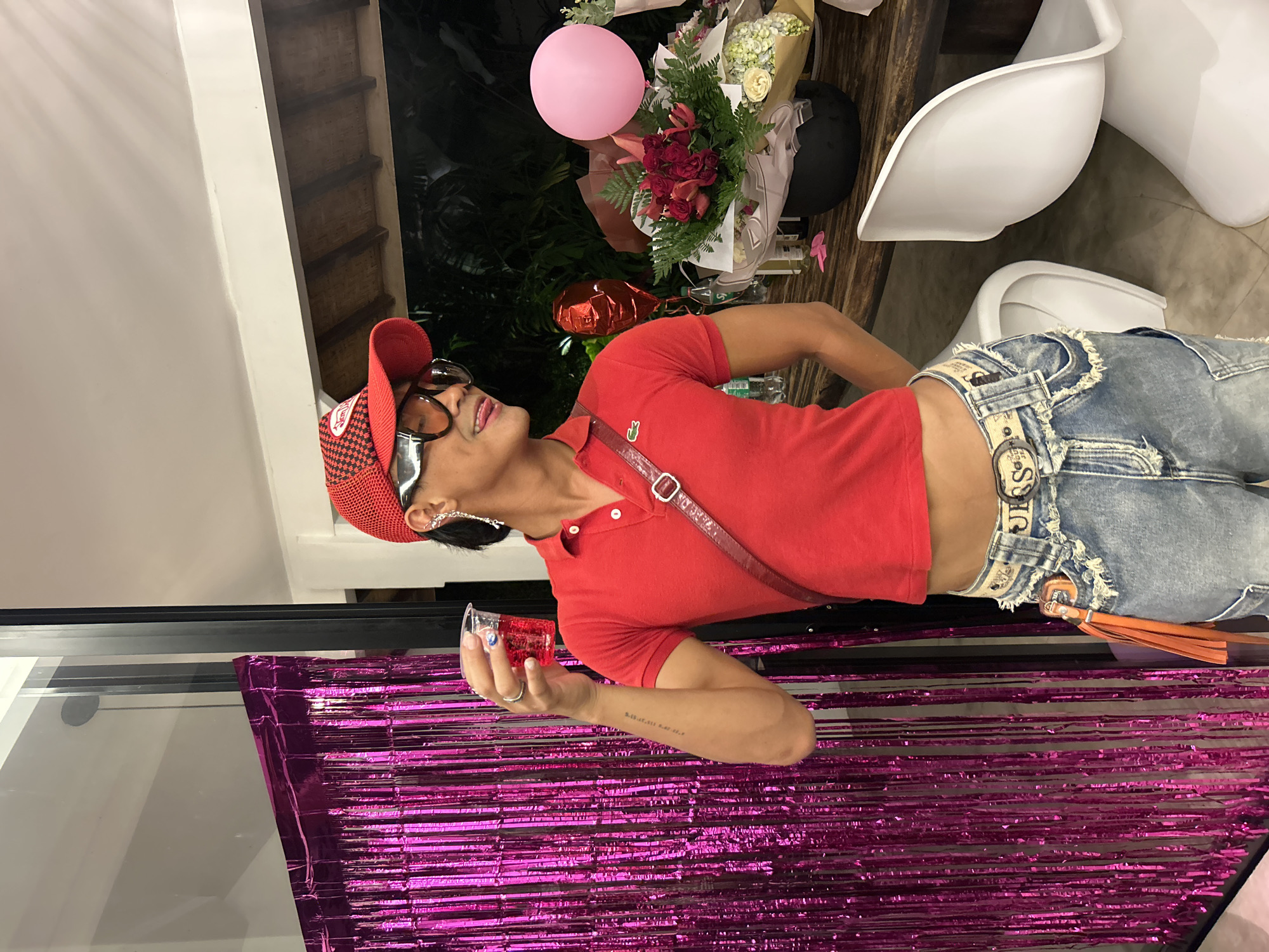 In North Kuta, Indonesia, a person wearing a red polo shirt, a red cap, and distressed denim jeans stands near a wall decorated with shiny pink fringe. They are holding a clear plastic cup filled with a red liquid and wearing large sunglasses and an ear accessory. In the background, a wooden table is laden with a bouquet of red and pink flowers, a pink balloon, and other unidentifiable items. Two white chairs are positioned near the table. The lighting suggests it is evening.