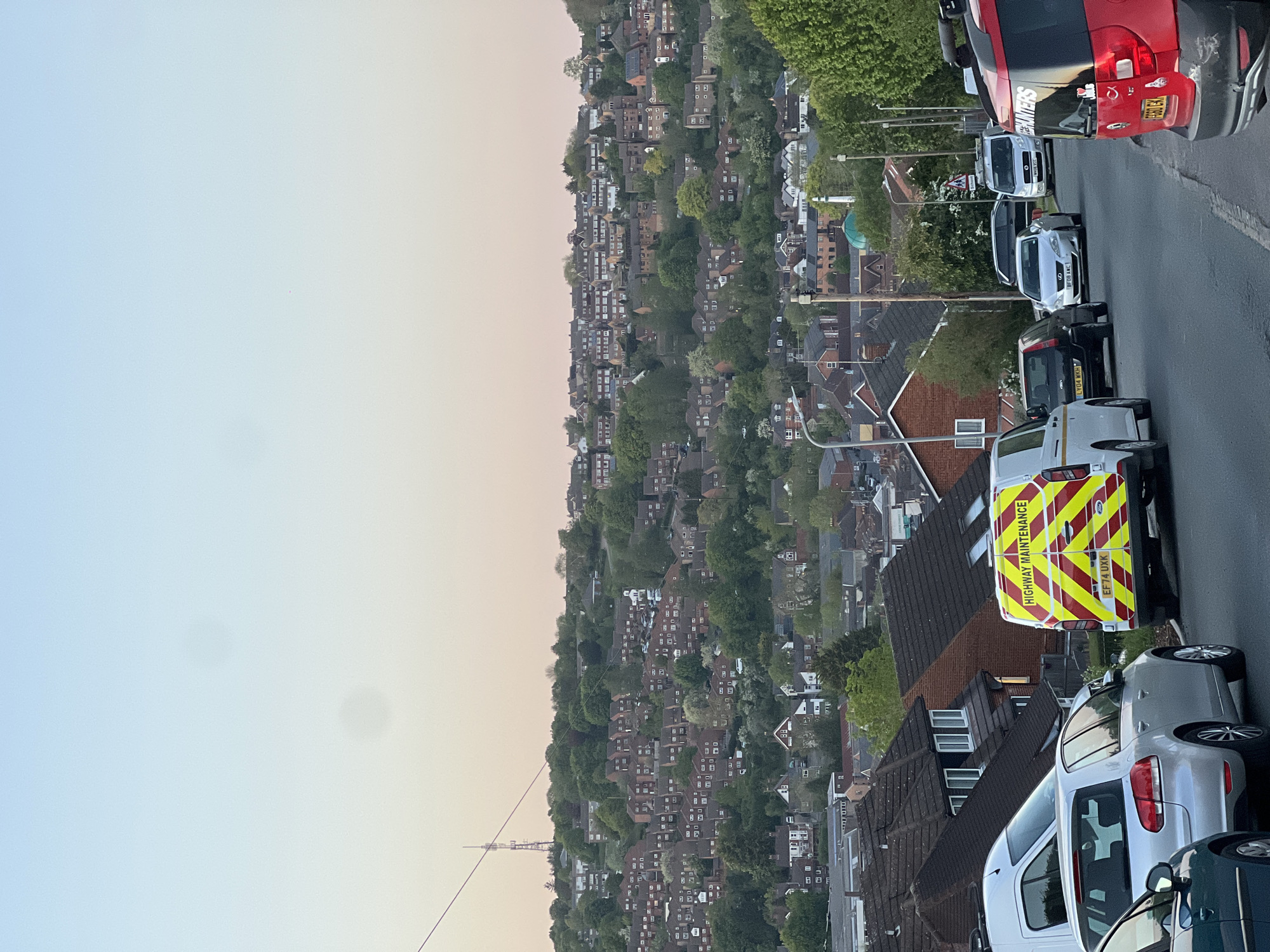 The image, originally oriented sideways, displays a panoramic view of a residential area built on a hillside, characteristic of towns like High Wycombe, United Kingdom. The time of day appears to be either sunrise or sunset, indicated by the soft gradient of the sky, which shifts from a pale blue on the left to a warm, light orange-pink hue on the right.
In the foreground, a paved street is visible, lined with several parked vehicles. Closest to the viewer is the rear of a white van with prominent yellow and red chevron markings and the text "HIGHWAY MAINTENANCE." Its license plate reads "EF71 UXX." Further along the street, a silver car (possibly a Volkswagen Golf variant) and a partially visible dark-colored car are parked. More vehicles are visible further down, including a black car with license plate "LT04 RNH," a white Ford Focus with license plate "WF08 LWC," another white van, and a red commercial van or truck. The red vehicle has partial text, possibly "PLUMBERS," on its side and a license plate "GJ10 EKA."
Beyond the street, the hillside is densely populated with numerous houses, featuring brick roofs and varied architectural styles, largely nestled among abundant green trees. A notable architectural feature, a dome-shaped building, can be seen among the houses on the right side of the hillside. In the far distance, towards the left horizon, a tall, slender mast or tower structure is faintly visible against the twilight sky. Streetlights and overhead utility cables are also scattered throughout the scene. The overall impression is a serene, well-established urban landscape integrated into a natural, hilly environment.