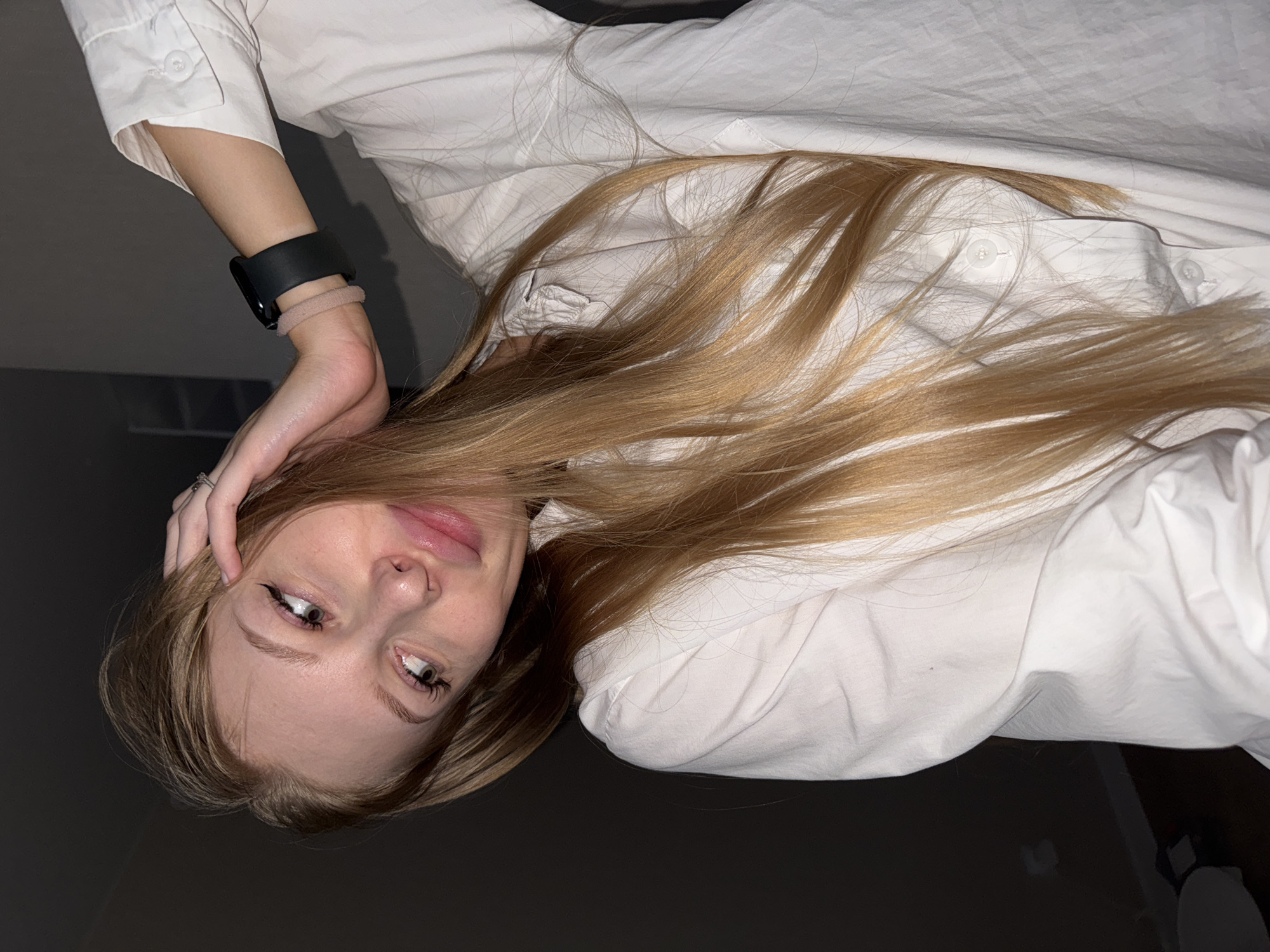 A young blonde woman with long hair, wearing a white button-down shirt and a black smartwatch, is in a dimly lit indoor setting. She is leaning her head to the side, with her left hand touching her hair near her temple. Her right arm is raised, with her hand resting behind her head. Her gaze is directed to her left, and her lips are slightly parted. The background appears to be a plain, dark wall. There is no indication of the city of Perm, Russia, in this image.