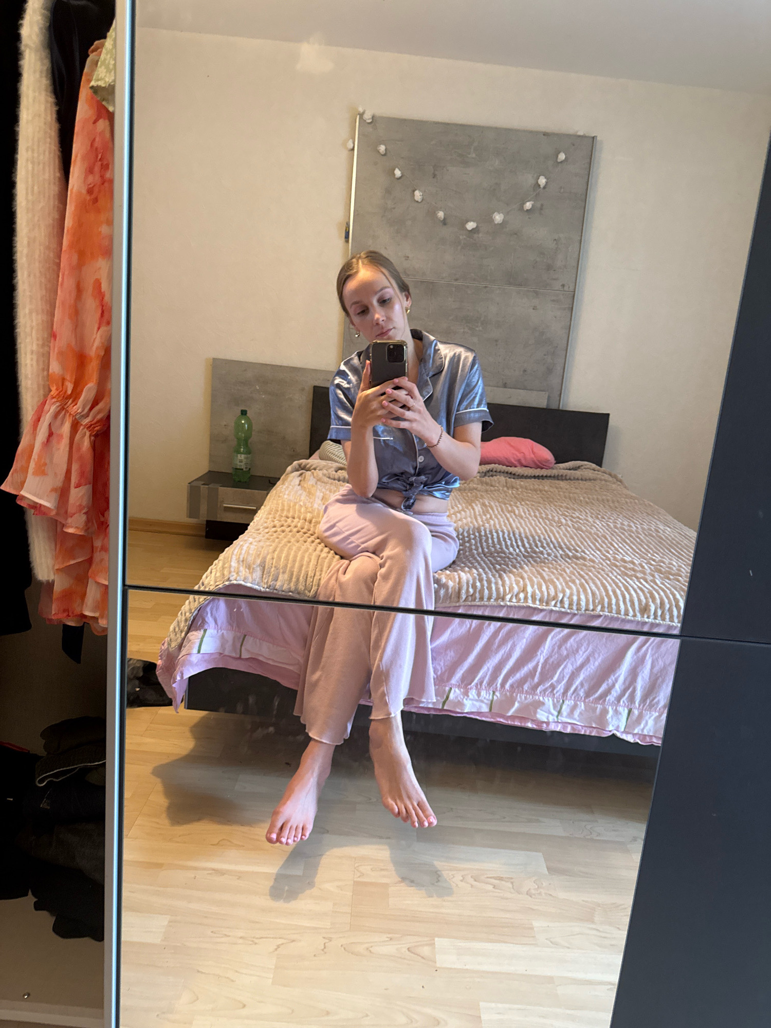 The image shows a full-length mirror reflecting a young woman sitting on a bed in a bedroom. She is wearing pajamas, a silk button-up top tied at the waist and loose-fitting pants. The woman is holding a smartphone in her right hand, taking a selfie. The bed has a textured beige comforter and a pink bedsheet. A bedside table with a green bottle on it is visible to the left of the bed. In the reflection, a closet with clothing is seen on the far left. The floor is light-colored wood. The wall behind the bed has a large, grey concrete panel with decorative elements. The room appears to be a bedroom in a residential apartment or house, likely located in the city of Überherrn, Germany.