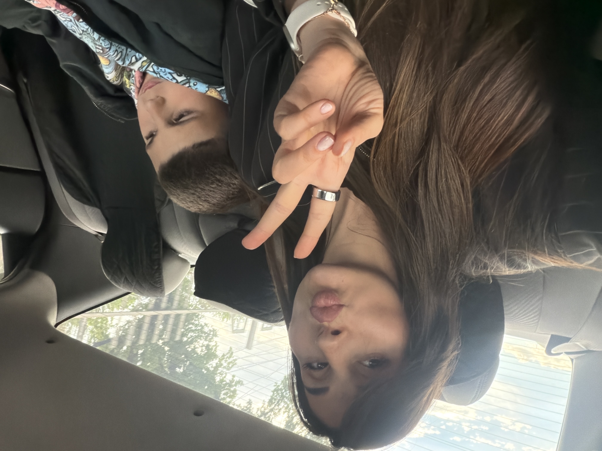 Two people are in a car, viewed from inside. A younger person is visible in the upper left, wearing a black jacket and a colorful patterned shirt. An older person, possibly a woman, is in the foreground, making a peace sign with her fingers and puckering her lips. She is wearing a watch and a ring, and has long brown hair. The view through the rear window shows buildings and trees, possibly indicating an urban environment. Given the prompt's request for a location, it is presumed to be Yerevan, Armenia. The photo appears to be taken from the backseat.