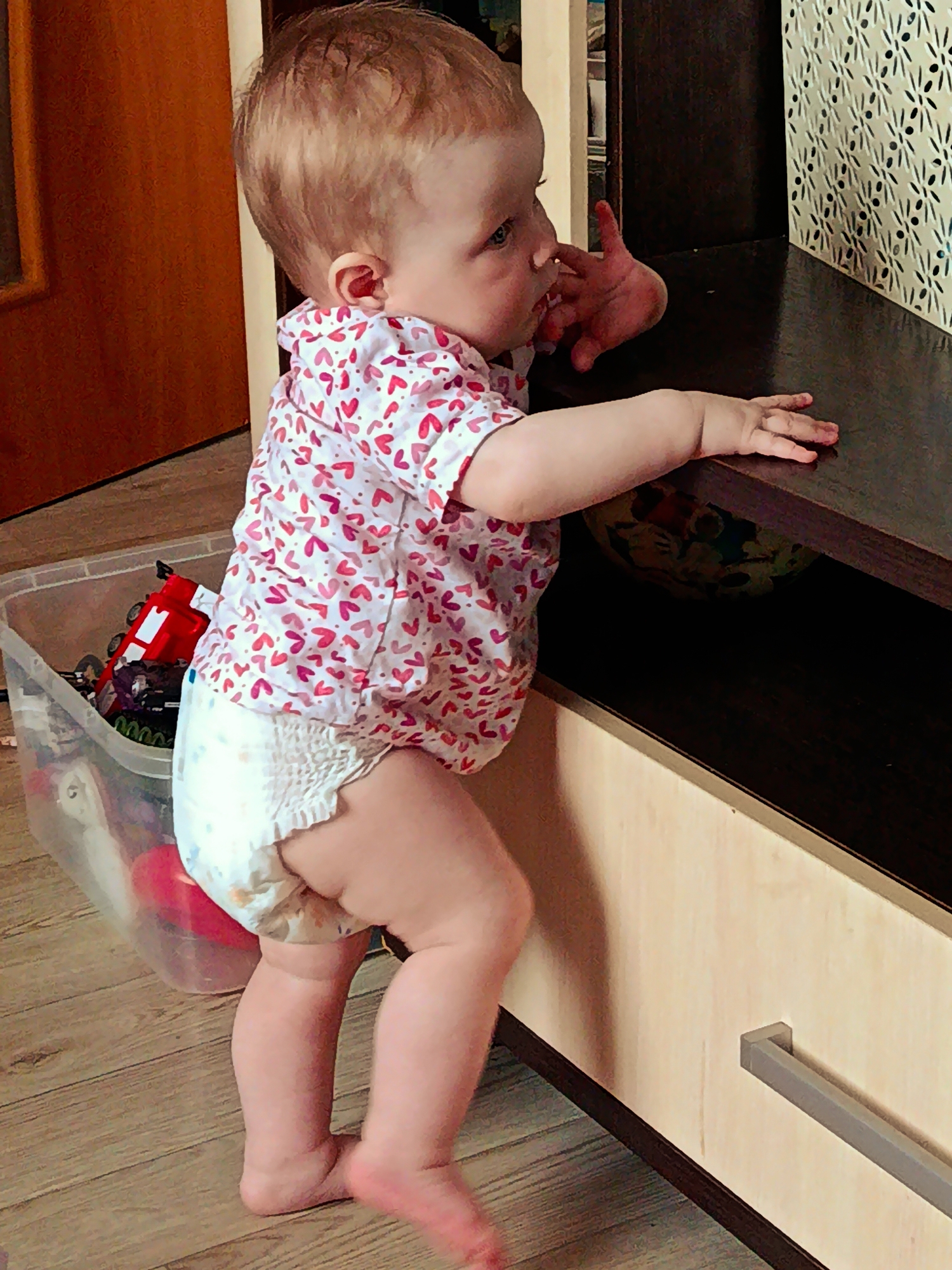 A toddler stands in front of a dark brown and beige cabinet with a silver handle. The child is wearing a diaper and a light pink and white shirt patterned with hearts. They are standing on a wooden floor, using one hand to support themself on the cabinet's surface. The toddler's other hand is raised to their mouth. Beside the child is a clear plastic storage container filled with toys, including a red truck. A wooden door is visible in the background. The location is a room, possibly in a house or apartment, in the city of Фоки, Russia.
