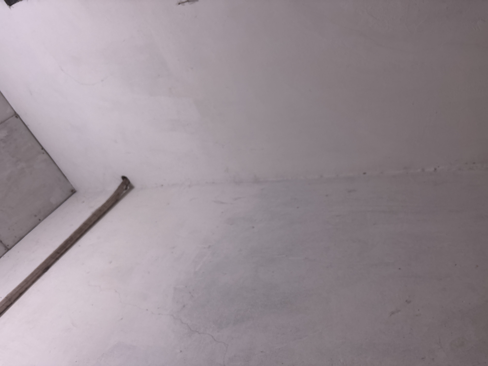 A dimly lit interior corner of a building shows two plain, off-white walls meeting at a right angle. A dark, narrow conduit or pipe runs diagonally across the upper section of the image, along the seam where the walls meet the ceiling. The ceiling itself is a light gray, concrete-like material. The walls appear textured and slightly uneven, with faint cracks and discoloration visible on the left wall. There are no people, animals, or any other objects or actions depicted in the scene. The location is an unadorned, unfinished space, possibly a utility area or a building under construction.
