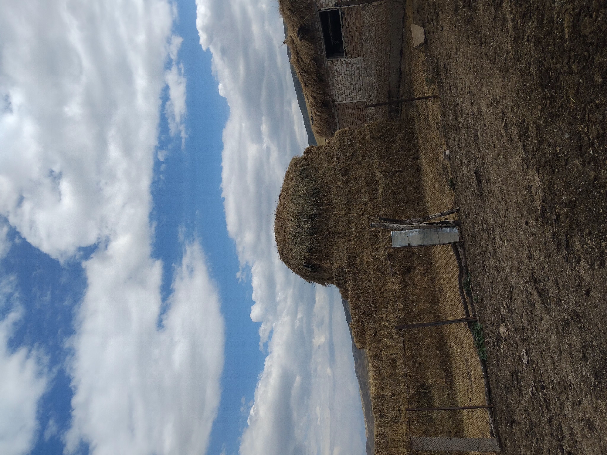 The image depicts a rural scene in Kopuro Bazar, Kyrgyzstan, characterized by a collection of hay bales and a simple structure under a cloudy sky. A large, conical stack of hay dominates the center of the frame, with smaller rectangular bales forming a lower wall to its left. Adjacent to the hay, a brick building with a thatched roof and a dark, open window is visible. The ground in the foreground is rough, dry earth, suggesting an agricultural or farm setting. The sky is a mix of blue and white clouds, indicating partly cloudy weather. No people are present in the image.