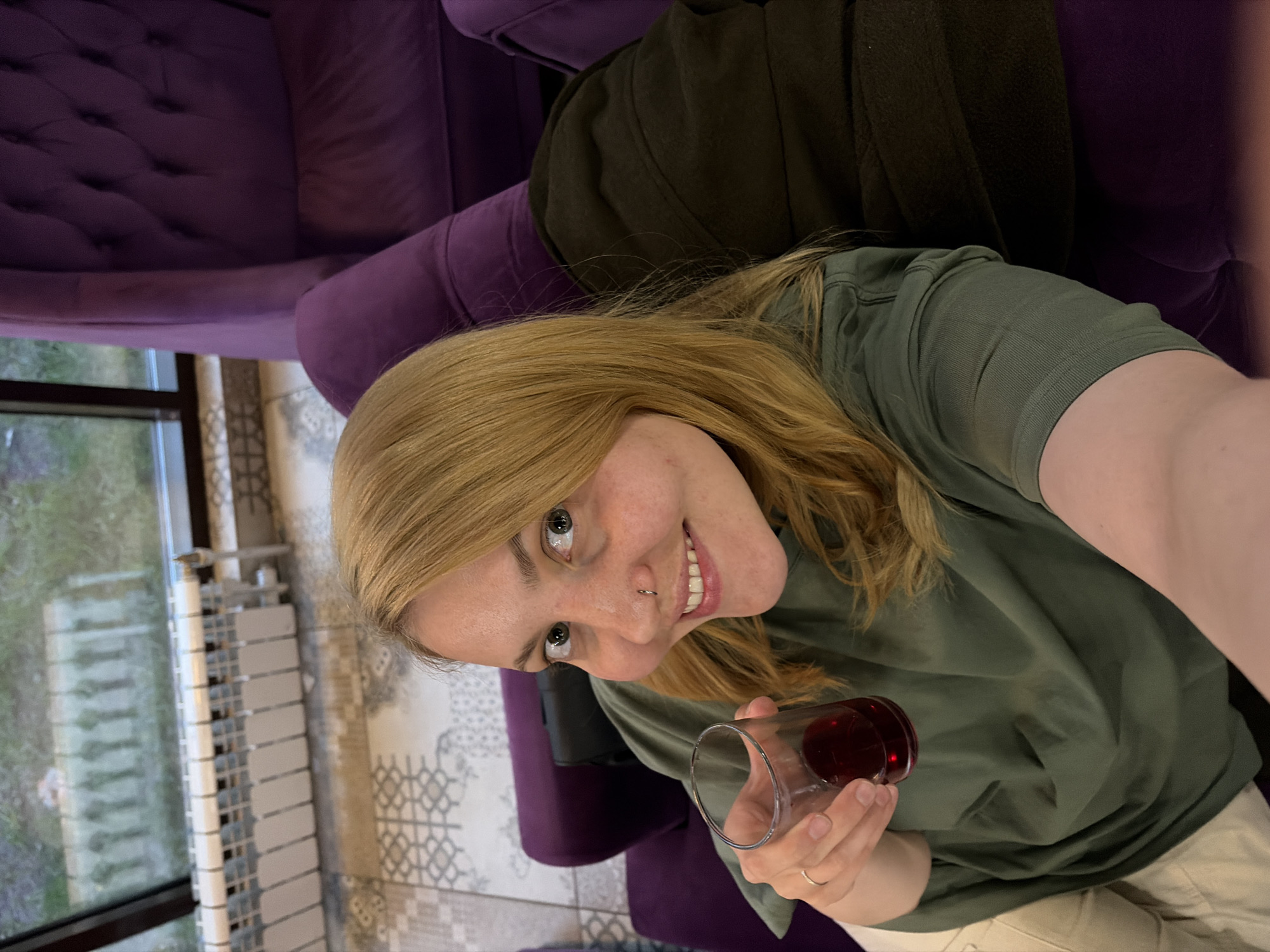 A woman with light blonde hair and a nose ring is holding a glass containing a red liquid. She is seated on a purple tufted couch, wearing an olive green t-shirt and light-colored pants. Behind the couch, there's a window with a view of trees. The floor is covered with a patterned tile. The location is the city of Chaikovsky, Russia.