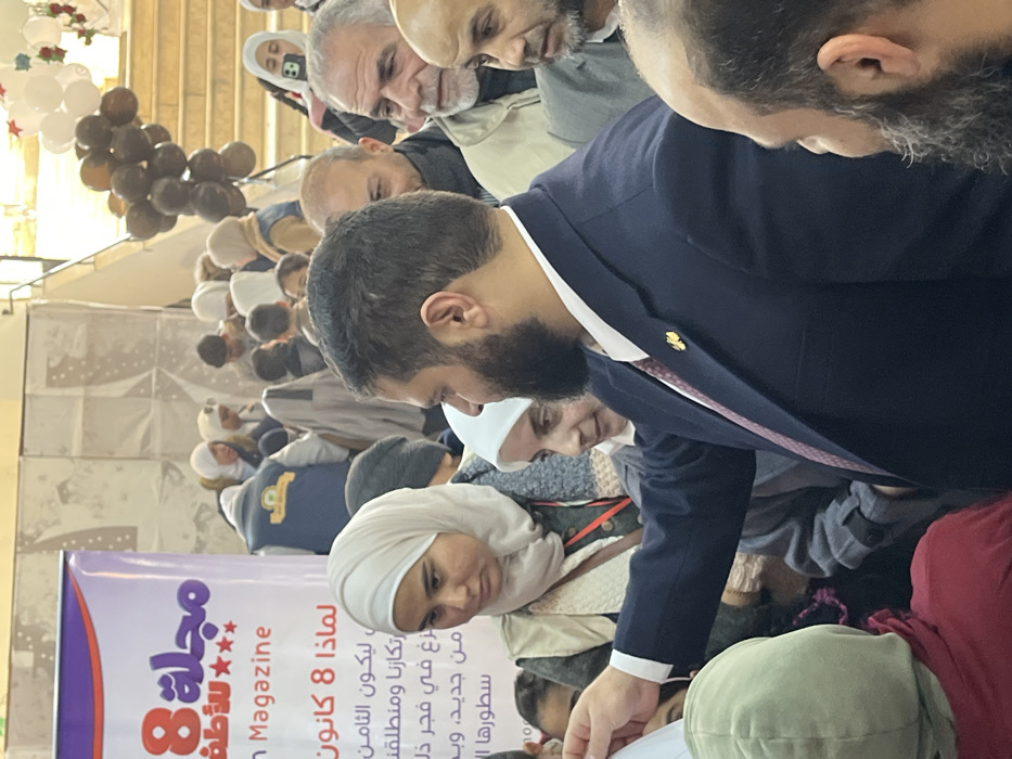 A densely populated indoor gathering is depicted in Kafr Batna, Syria. In the foreground, a male subject with a dark beard and dark hair, wearing a dark blue suit jacket, a white shirt, and a burgundy tie with a gold lapel pin, extends his right arm forward, appearing to interact with an individual partially obscured below the frame. To his left, a female subject wearing a light beige hijab and a light-colored vest over a dark garment looks downward and to her left. Behind her, another female subject with a white hijab is observed smiling.
The midground and background are populated by numerous individuals, including adults and children, many wearing head coverings (hijabs) and various winter clothing items. On the far left, a large banner is partially visible, displaying purple and red text in Arabic and English, including "Magazine 8 for Children" and "Why January 8." In the upper right background, a light-colored staircase is present, adorned with clusters of dark brown and white balloons, and star-shaped decorations. The overall environment suggests a public event or celebration within a brightly lit indoor space.