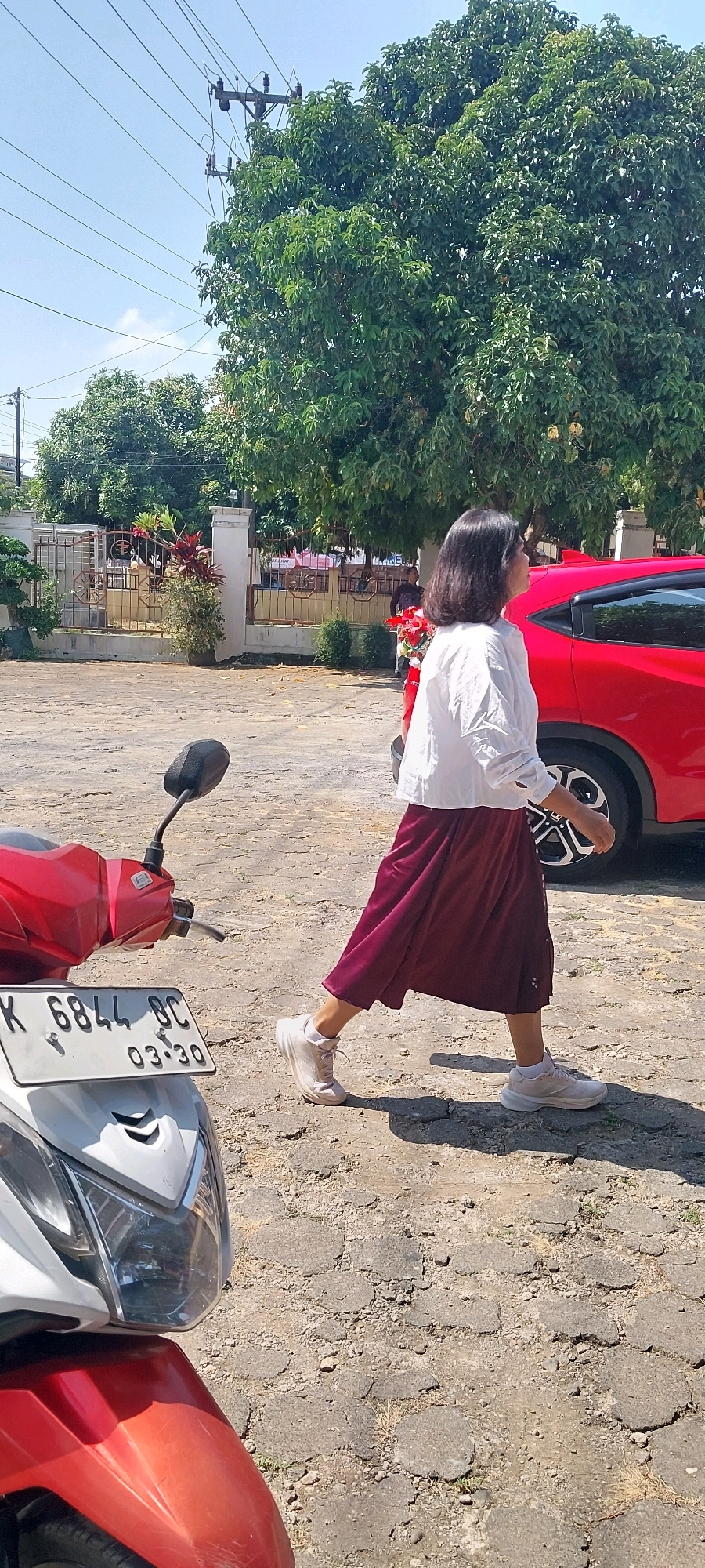 In Keling, Indonesia, a woman walks in a paved outdoor area. She is wearing a white blouse and a maroon skirt, with light-colored sneakers. She is walking towards the right of the frame, with her back to the viewer. In the foreground, the front of a red and white motorcycle is visible. To her right, a red car is parked. The background includes a fence, trees, and utility poles with wires. The ground is rough and cracked pavement. The sky is clear and blue.