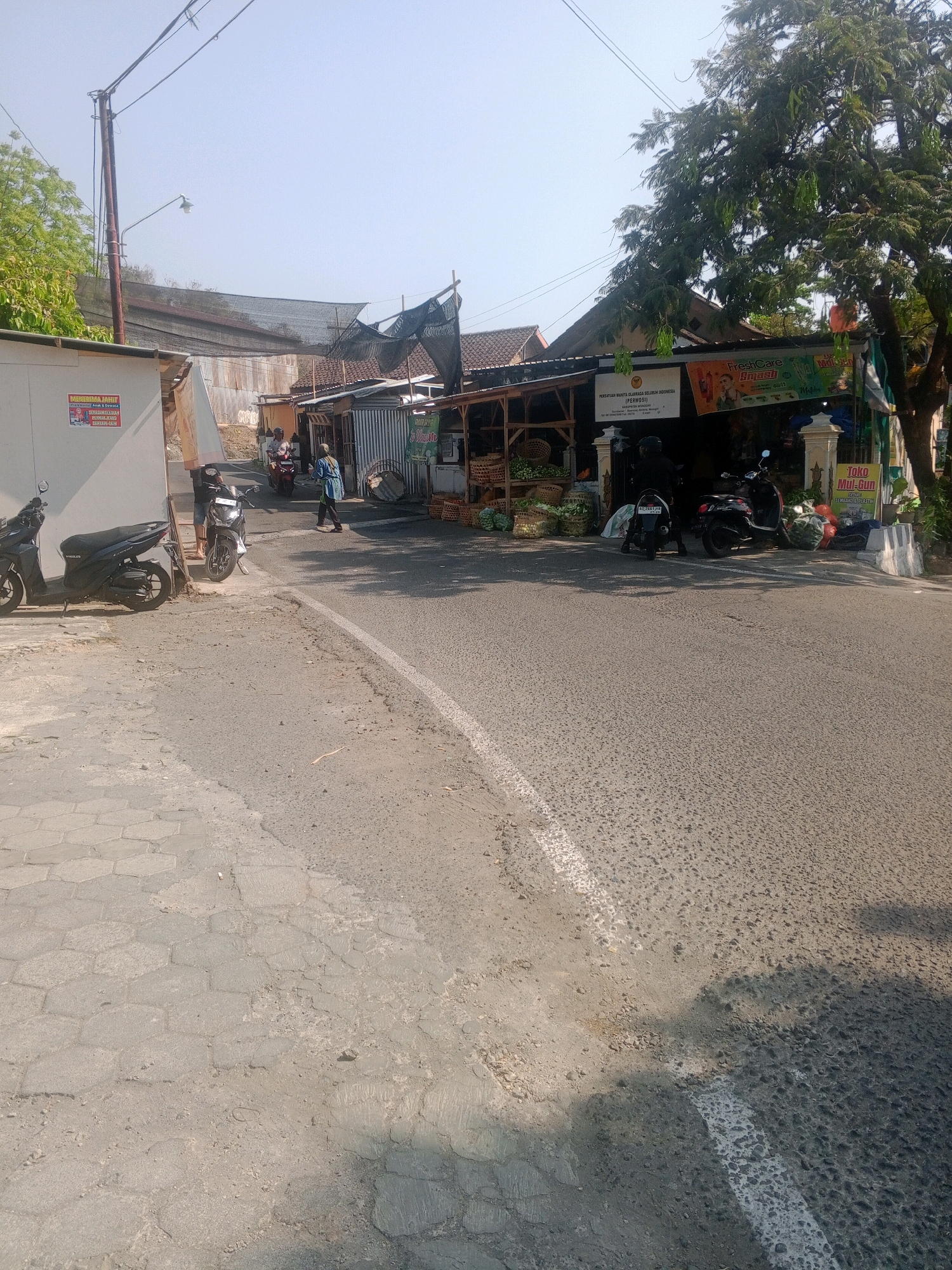 This is an eye-level, slightly high-angle, full shot taken outdoors on a sunny day in what appears to be a commercial area on the outskirts of Wonogiri, Indonesia. The scene depicts a street with parked motorcycles and a small market stall. On the left side of the image, a white building with a sign that reads "MENERIMA JAHIT" (Accepting Sewing) is visible. Several parked motorcycles, including a dark-colored scooter and a lighter-colored one, are positioned in front of the building.
In the center and right of the image, a market stall with a corrugated metal roof and wooden shelves is set up. The stall displays various goods, including what appear to be vegetables in baskets and bags. A sign above the stall reads "FreshCare smash" and a smaller sign to its right says "Toko Mul-Gun" (Mul-Gun Shop) with the text "SEMBAKO & PASTA" (Groceries & Pasta). Two people are visible near the market stall; one person in a blue long-sleeved shirt and light-colored pants is walking away from the stall, and another person in a black outfit and helmet is standing near a parked motorcycle in front of the stall. Another motorcycle is parked further back and a person wearing a helmet is riding it. A large tree with hanging pods partially obscures the market stall and the sky. The street is paved with asphalt and features a white line marking. The sidewalk on the left side is made of hexagonal paving stones. The background shows houses and some vegetation. The overall impression is a typical roadside market scene in a town or rural area of Indonesia.