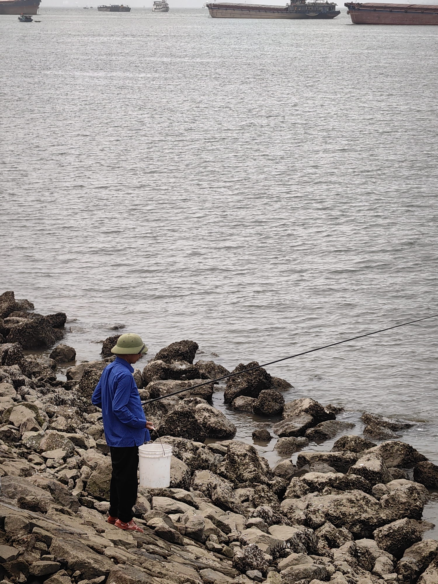 An eye-level medium shot shows a man fishing from a rocky coastline in Hong Gai Ward, Vietnam. The man, wearing a blue long-sleeved shirt, black pants, red sandals, and a green conical hat, stands with his back to the viewer. He holds a white bucket and extends a fishing rod towards the water. The coastline is comprised of various-sized rocks, many covered with what appears to be marine growth. The water is gray and choppy, with several large cargo ships visible in the distance under an overcast sky.