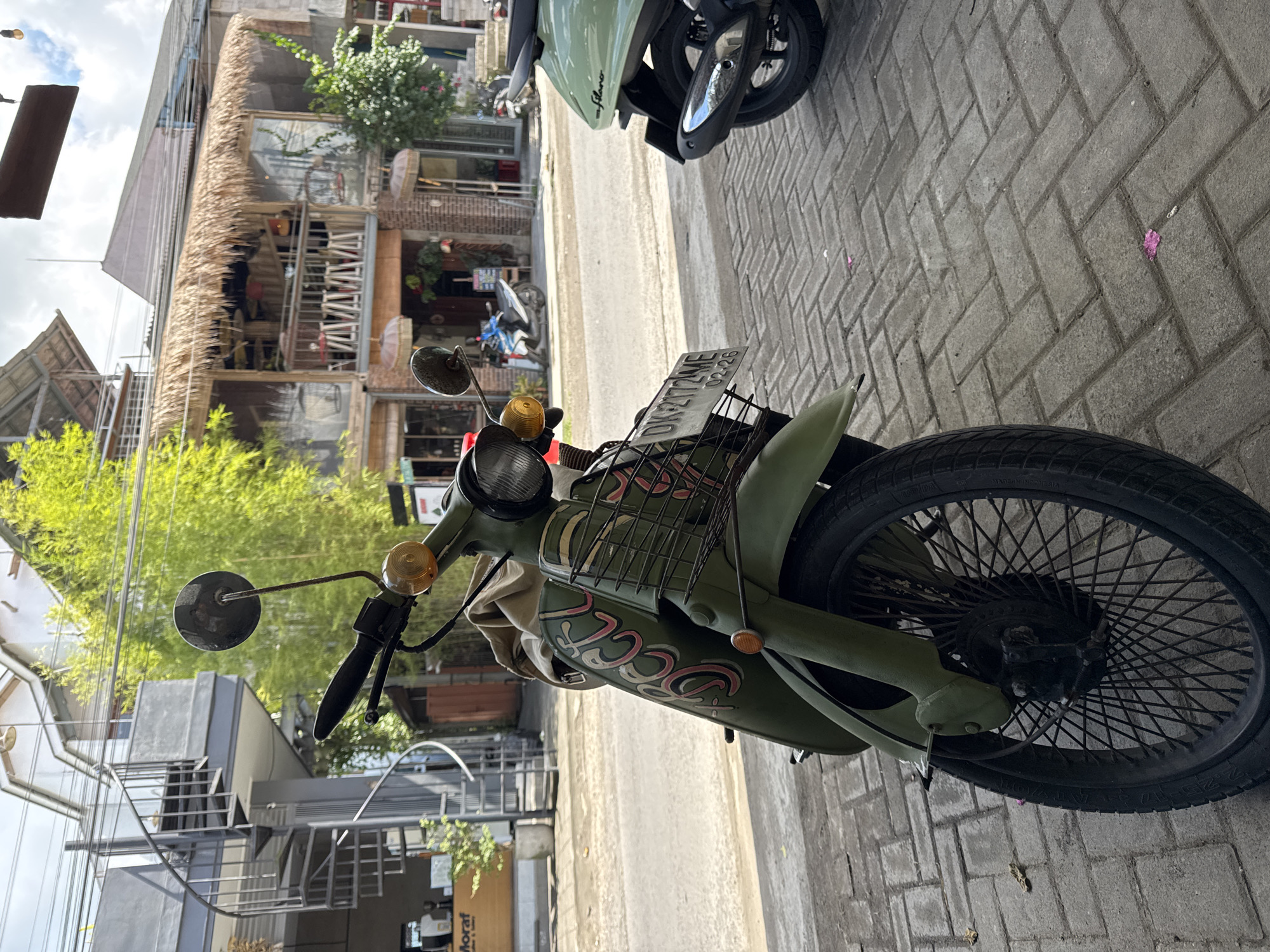 The image captures a street scene, likely in Kediri, Indonesia, as indicated by the context. The foreground is dominated by a vintage-style green scooter, parked on a grey paved sidewalk. The scooter features a prominent round headlight, amber turn signals, side mirrors, and a metal basket mounted at the front. A license plate, bearing the text "DK 2172 WE" and "02-26", is visible above the basket. The side of the scooter displays a stylized pinkish-red graphic that appears to spell "SCOO". Its front wheel has numerous black spokes, and the tire shows "MADE IN INDONESIA".
Behind the green scooter, another light green scooter is partially visible on the sidewalk. A paved road stretches between the sidewalk and a row of buildings. These buildings exhibit a rustic or traditional architectural style, with some featuring distinctive thatched roofs and exposed brickwork. They appear to be multi-story structures, possibly housing cafes or shops, with visible windows and open sections. Lush green foliage, including bamboo-like plants, grows around and behind the buildings, adding a natural element to the urban environment. The sky above is bright and partly cloudy, indicating daytime. A small sign on one of the background buildings mentions "Moraf". The overall scene suggests a vibrant local setting with a mix of traditional and modern elements.