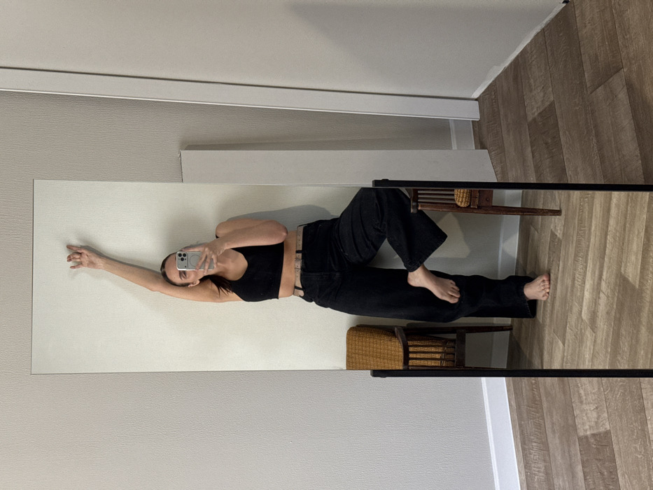 The scene depicts a female individual in the reflection of a full-length, black-framed mirror, located indoors in the city of Chaikovsky, Russia. The individual, presumed to be an adult, is positioned centrally within the mirror's view, facing the mirror to capture a selfie with a light-colored smartphone held in her right hand, which partially obscures her face. Her dark hair is pulled back. She is dressed in a black sleeveless crop top and loose-fitting black trousers with a light-colored belt. Her left arm is extended upwards, with the hand resting against the light-colored, textured wall behind her. Her right leg is bent, with her bare right foot resting on the patterned, possibly upholstered, seat of a dark wooden chair. Her left foot is bare and rests on the light brown laminate flooring laid in a staggered plank pattern. A second, similar dark wooden chair is partially visible to the left background of the reflection, further back in the room. The immediate environment includes a white baseboard, a light-colored wall, and a white vertical panel or door frame visible on the right side of the reflection.