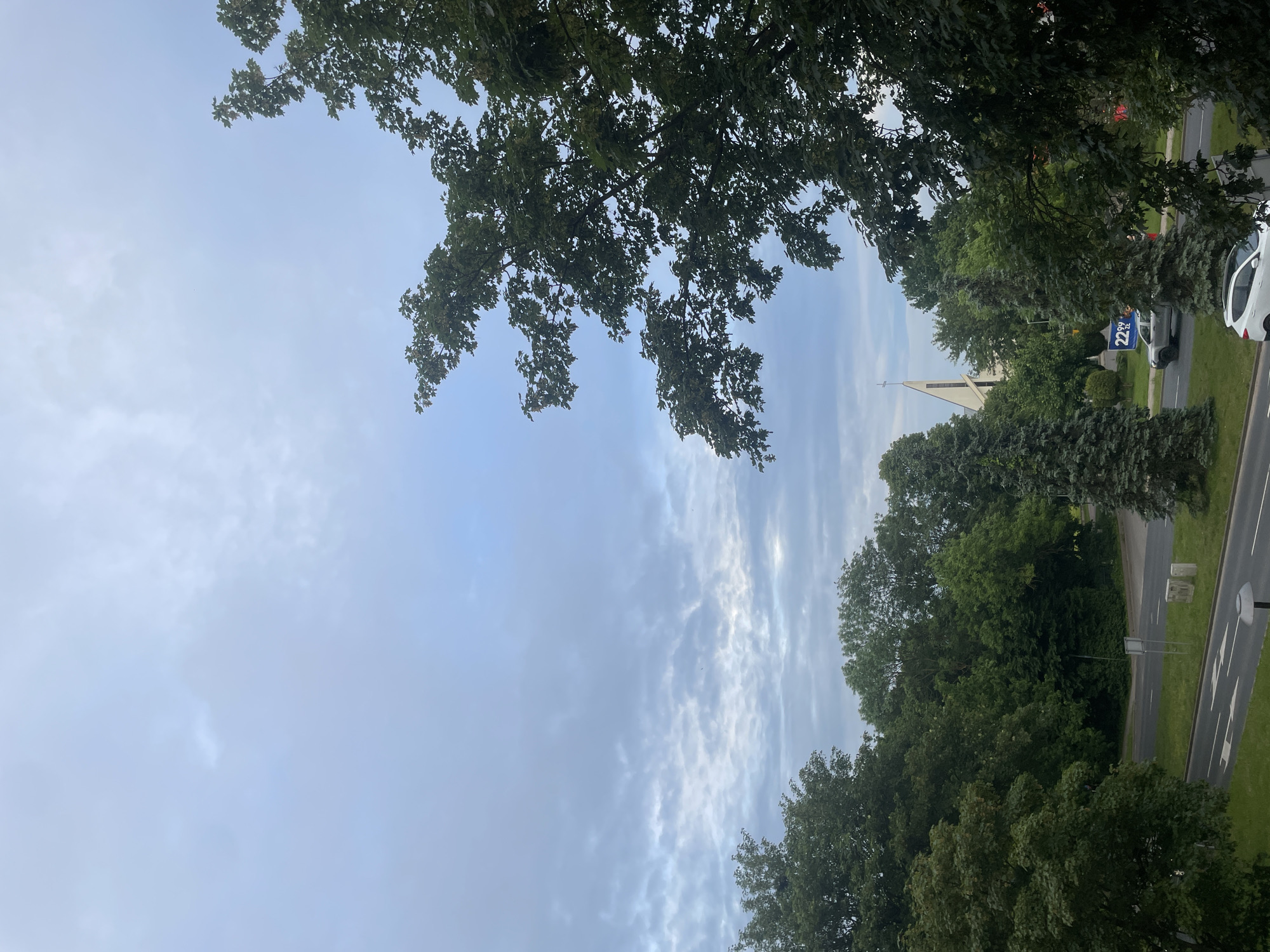 This is a slightly overcast, daylight shot of an urban scene in Kolobrzeg, Poland. The foreground is dominated by the dense green foliage of mature trees on the right and left, with branches extending into the frame and obscuring parts of the sky. A paved road with white lane markings and directional arrows runs diagonally across the lower portion of the image. Two cars are visible on the road: a white SUV in the lower right corner and a silver SUV further back, to the left. A church with a tall, pointed steeple is visible in the mid-ground, situated behind a line of trees and a blue banner with white text. The sky is filled with a mixture of light and dark clouds. The overall impression is one of a residential or commercial area with significant tree cover.
