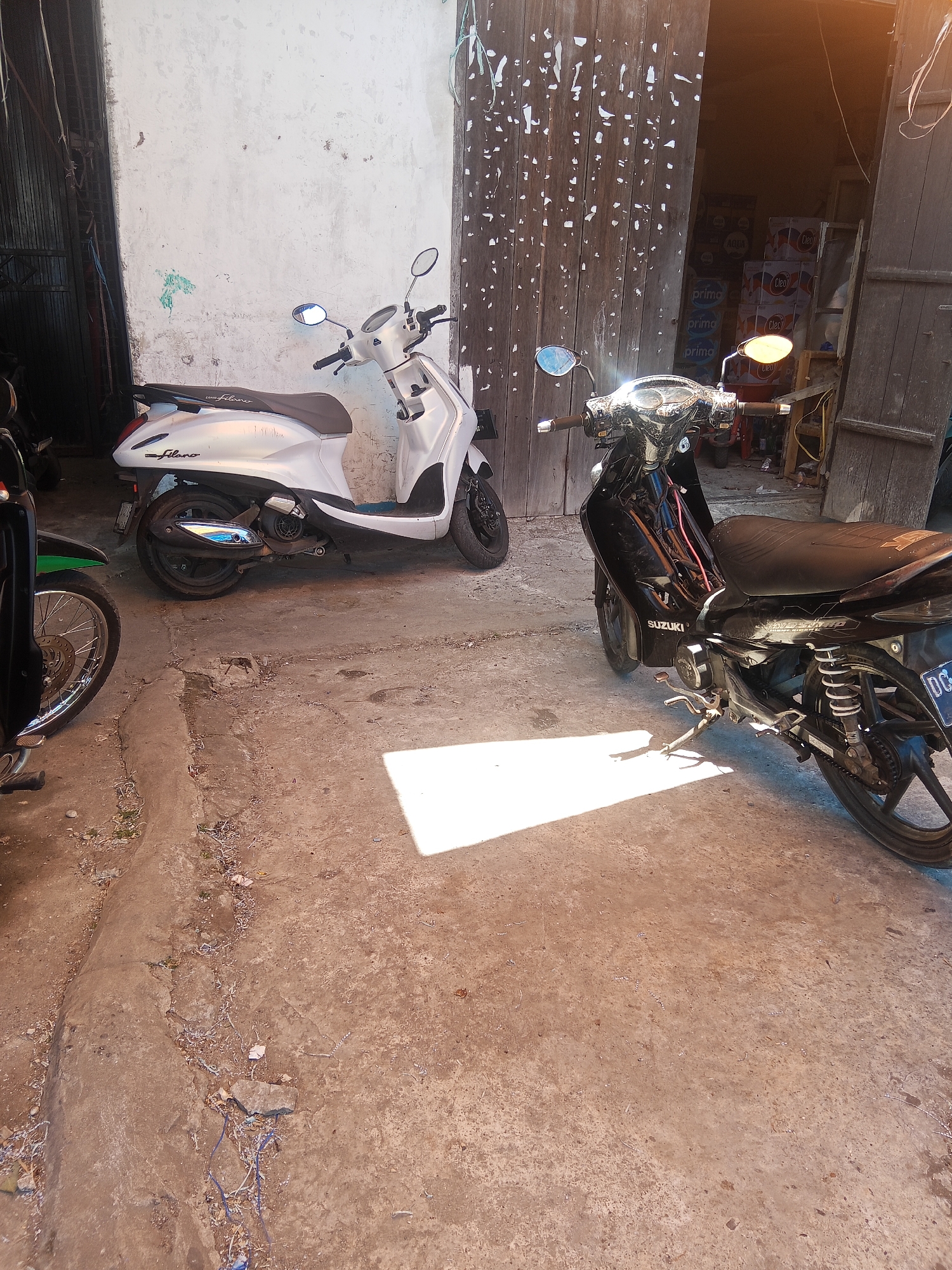 Two motorcycles are parked in front of a weathered wooden door, with a white wall to the left. The motorcycle on the left is white and silver with the brand name "filano" visible on its side. It has a black seat and a chrome exhaust pipe. The motorcycle on the right is black and appears to be a Suzuki model, with visible wiring and a black seat. Its license plate is partially obscured but shows "DCS". The ground is concrete and appears worn, with scattered debris. In the background, through the open wooden door, stacks of bottled water are visible. The lighting suggests it is daytime, with a strong sunlight casting shadows on the ground. The scene is likely an alleyway or a service entrance in Makale, Indonesia, a city in the Toraja region known for its distinctive traditional architecture, though this particular location appears more utilitarian.
