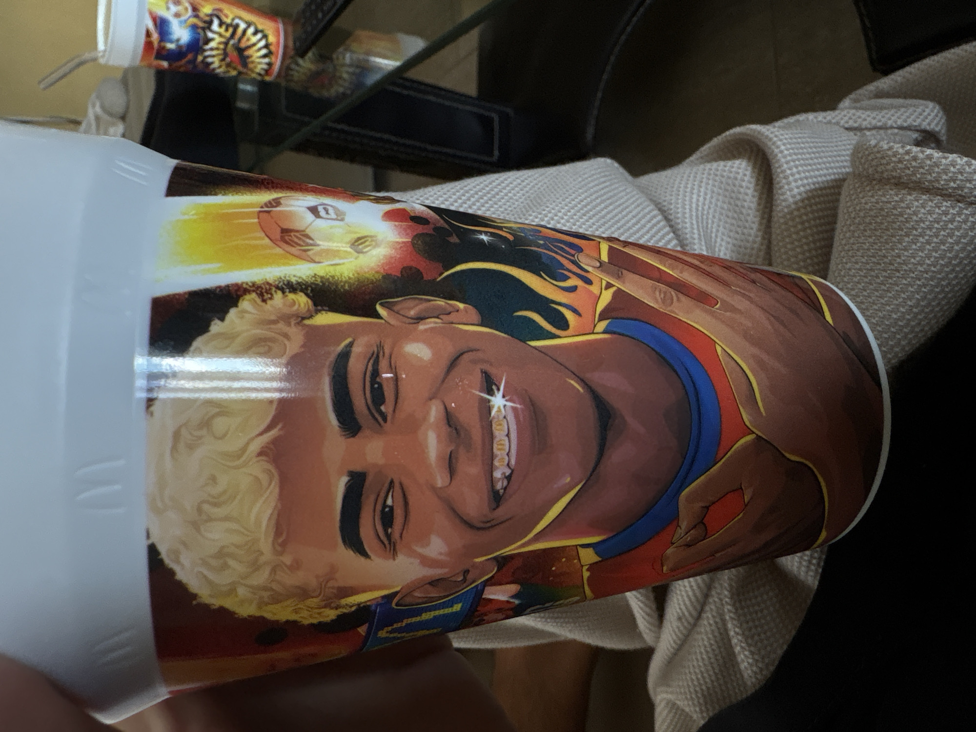 The image shows a white plastic cup with a vibrant graphic wrapping around it. The graphic features an illustration of a young person with curly blonde hair and dark skin, wearing a red and blue outfit. They have a wide smile, revealing braces that are sparkling. Their left hand is raised, with fingers forming a circle, and a starburst is visible near their mouth. The background of the illustration shows a soccer ball in motion against a fiery, yellowish-orange backdrop, suggesting a theme related to sports or energy.
To the left of the cup, a small portion of another cylindrical object with a similar colorful graphic is visible. This object also appears to be a container, possibly another cup or a small bottle.
The cup is resting on a textured, light-colored fabric, which appears to be a blanket or a towel. The background is dark, with some hints of furniture or other objects, possibly in a room. The lighting seems to be indoor, as indicated by the general tone and the reflection on the cup's surface. The overall mood of the illustration on the cup is energetic and joyful. No discernible text is visible on the main cup, but the smaller object in the background might have some lettering on its graphic.