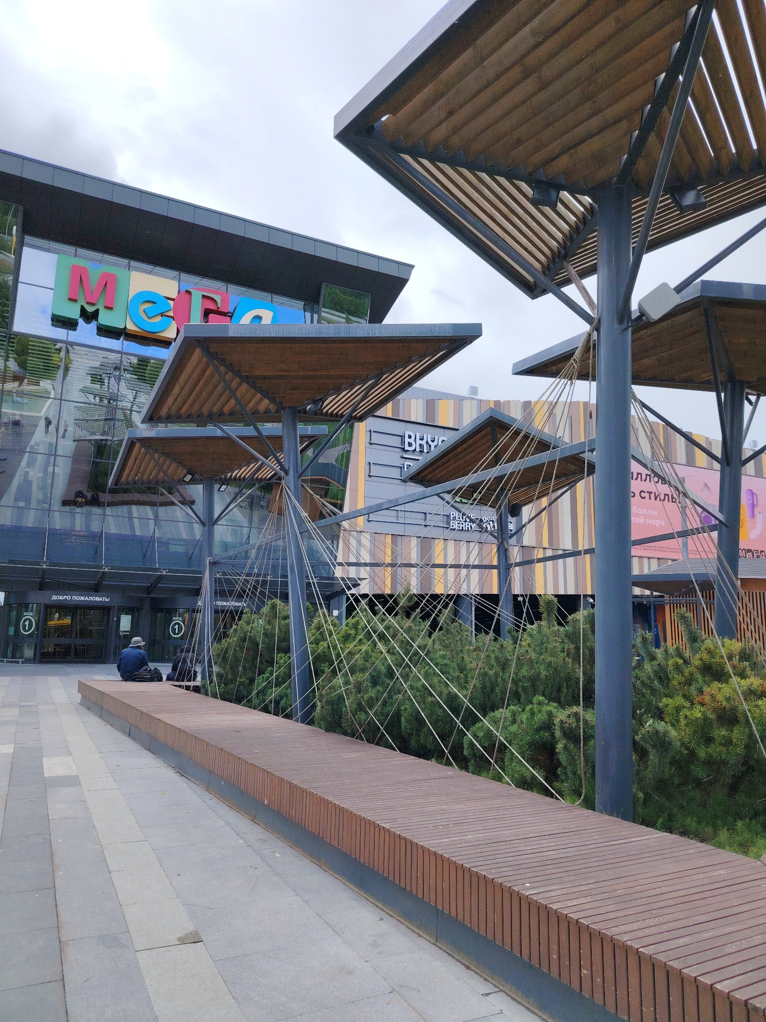 The image shows the entrance to the "METRO" hypermarket in the city of Yekaterinburg, Russia. A person is sitting on a long wooden bench near the entrance. There are architectural elements resembling stylized trees with wooden canopies supported by metal poles. Green bushes are planted around the structures. The hypermarket's entrance is visible with glass doors and a sign that reads "ДОБРО ПОЖАЛОВАТЬ" (Welcome). In the background, a building with vertical stripes and advertisements is visible. The sky is overcast.