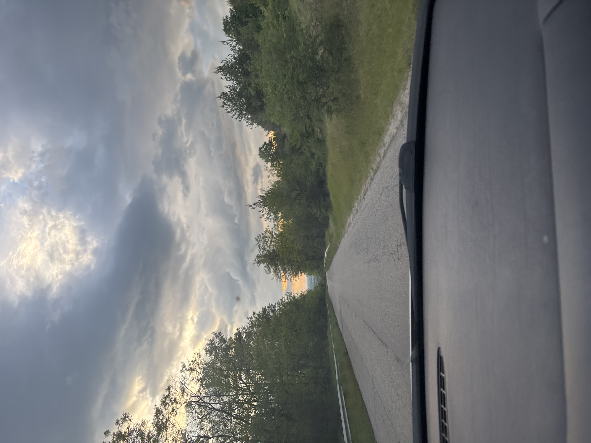 View from inside a car driving on a road. The road is asphalt with a centerline. Trees line both sides of the road. The sky is cloudy with some sunlight visible. Location is unknown.