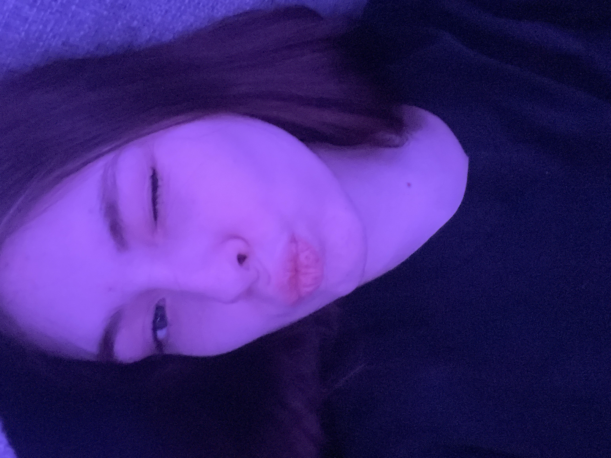 The image shows a close-up of a young person's face, likely a female, with one eye partially closed in what appears to be a wink or a squint. The skin appears pale, and the lighting casts a strong purple hue over the entire scene, altering the natural colors of the subject. The person's lips are slightly puckered. The subject's hair is dark, and a dark garment is visible around the neck and shoulder area. The overall scene gives the impression of being in a dimly lit indoor environment. There is no indication of this image being related to the city of Saint Petersburg, Russia.