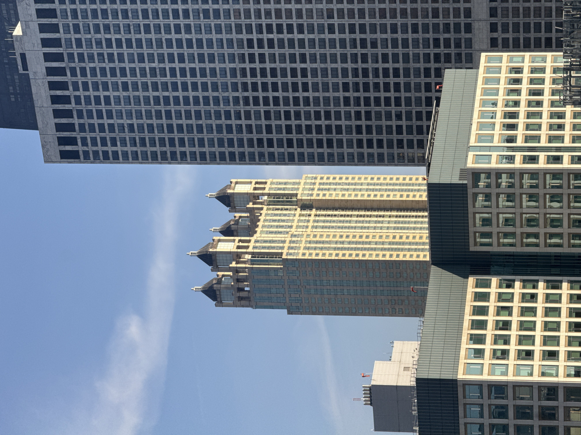 The image is an upward-facing shot of a collection of modern skyscrapers in Chicago. The sky is a clear, bright blue with a few wispy clouds, suggesting a sunny day, likely daytime given the illumination on the buildings. The architecture is characterized by geometric designs, with repeating patterns of windows and structural elements.
The most prominent building in the foreground is a light-colored skyscraper with a grid of square windows, many reflecting the blue sky. To its left, and slightly behind, is a darker, more massive building with a dense pattern of smaller, rectangular windows. The building in the center has a unique tiered design, with each level set back further than the one below, creating a layered effect with a yellowish facade and darker window frames.
The overall scene is one of urban density and architectural modernity. The sharp lines and vast scale of the buildings convey a sense of a bustling metropolis. There are no people visible, and the focus is entirely on the structures themselves and their interplay with the sky. The composition, with the buildings reaching upwards into the sky, emphasizes their height and grandeur. There is no visible text in the image.