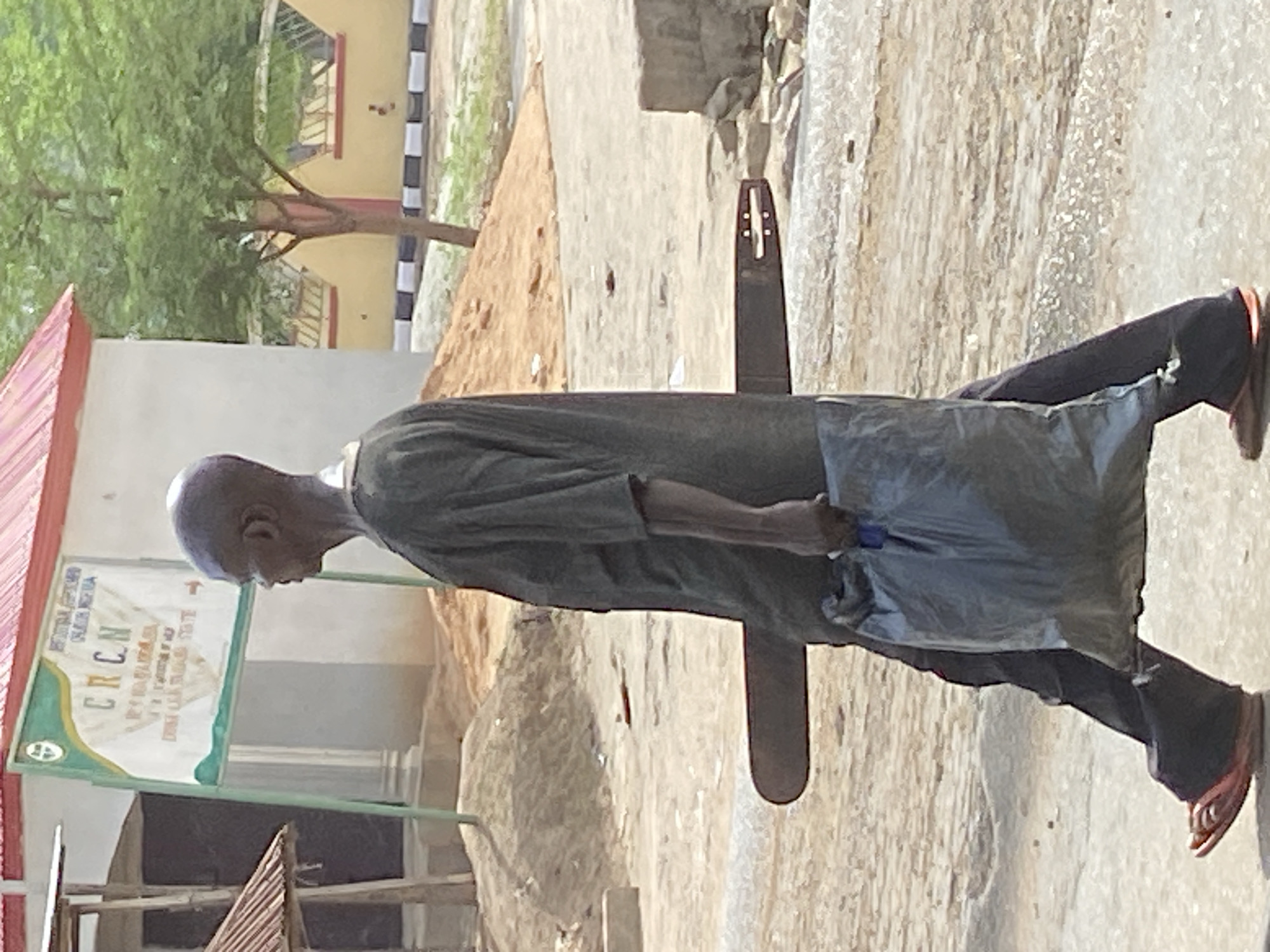 The image shows an elderly man walking on a paved area in Mararaba, Nigeria. He is wearing a dark shirt and pants with sandals. He carries a grey plastic bag. Behind him, there is a sign with the acronym "CRCN" on it, and a building with a red roof. A tree is visible in the background next to a building with a black and white striped accent. A dark brown, flat, object resembling part of a musical instrument rests next to the walking man.