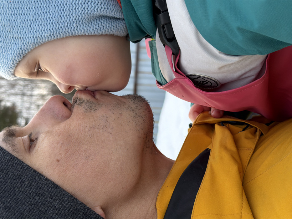 A close-up, side-profile image depicts an adult male and a child kissing. The adult male, on the left, wears a dark grey knit beanie and a bright yellow and black jacket, exhibiting light facial stubble. The child, on the right, wears a light blue knit beanie and a multi-colored jacket featuring teal, pink, and white panels. A black buckle and a circular metallic patch, potentially marked "DB" and "96," are visible on the child's jacket. Both individuals' lips are pressed together. The adult male's left hand is visible near the child's jacket. The background is blurred, showing a snow-covered ground, a light-colored corrugated fence, and bare trees, consistent with an outdoor winter setting. The precise location within the city of Chaikovsky, Russia, is not discernible from the image content.
