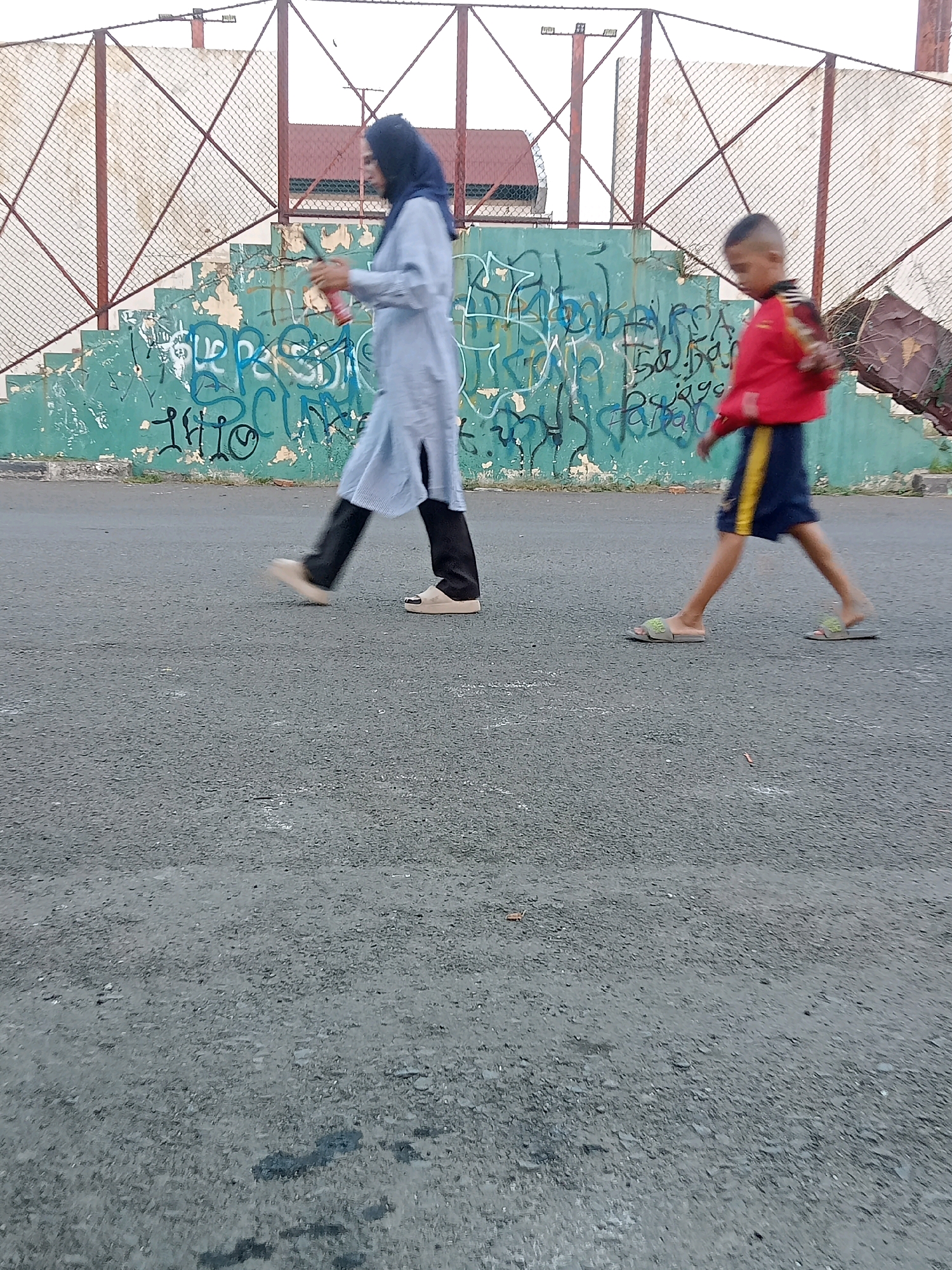 A woman wearing a light blue hijab and a light blue long-sleeved dress is walking from left to right, holding a red cylindrical object. A young boy in a red jacket and dark blue shorts with a yellow stripe is walking in the same direction to her right. They are on a paved surface in front of a green wall covered in graffiti, which is part of a set of stairs. A metal fence is visible behind the graffiti-covered wall. The scene appears to be outdoors in an urban area. The location is identified as Karawang, Indonesia.