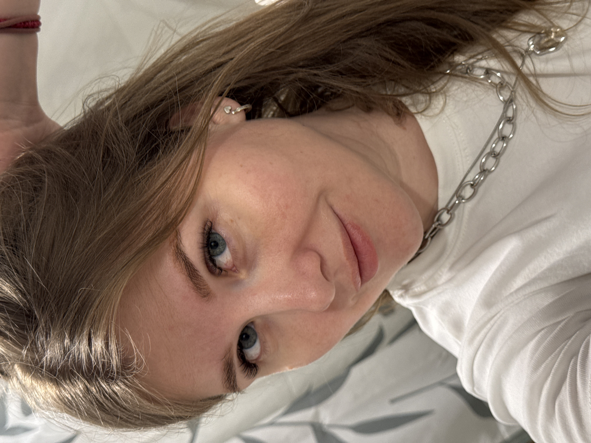 A close-up, angled portrait of a young woman with light brown hair and blue eyes is presented. She wears a white long-sleeved shirt and a silver chain necklace. A small earring is visible in her left ear. The background consists of a light-colored surface with a subtle pattern, possibly a bed sheet. There is no explicit information to connect this image to Gornyi (Dvurechenskoe S/P), Russia.