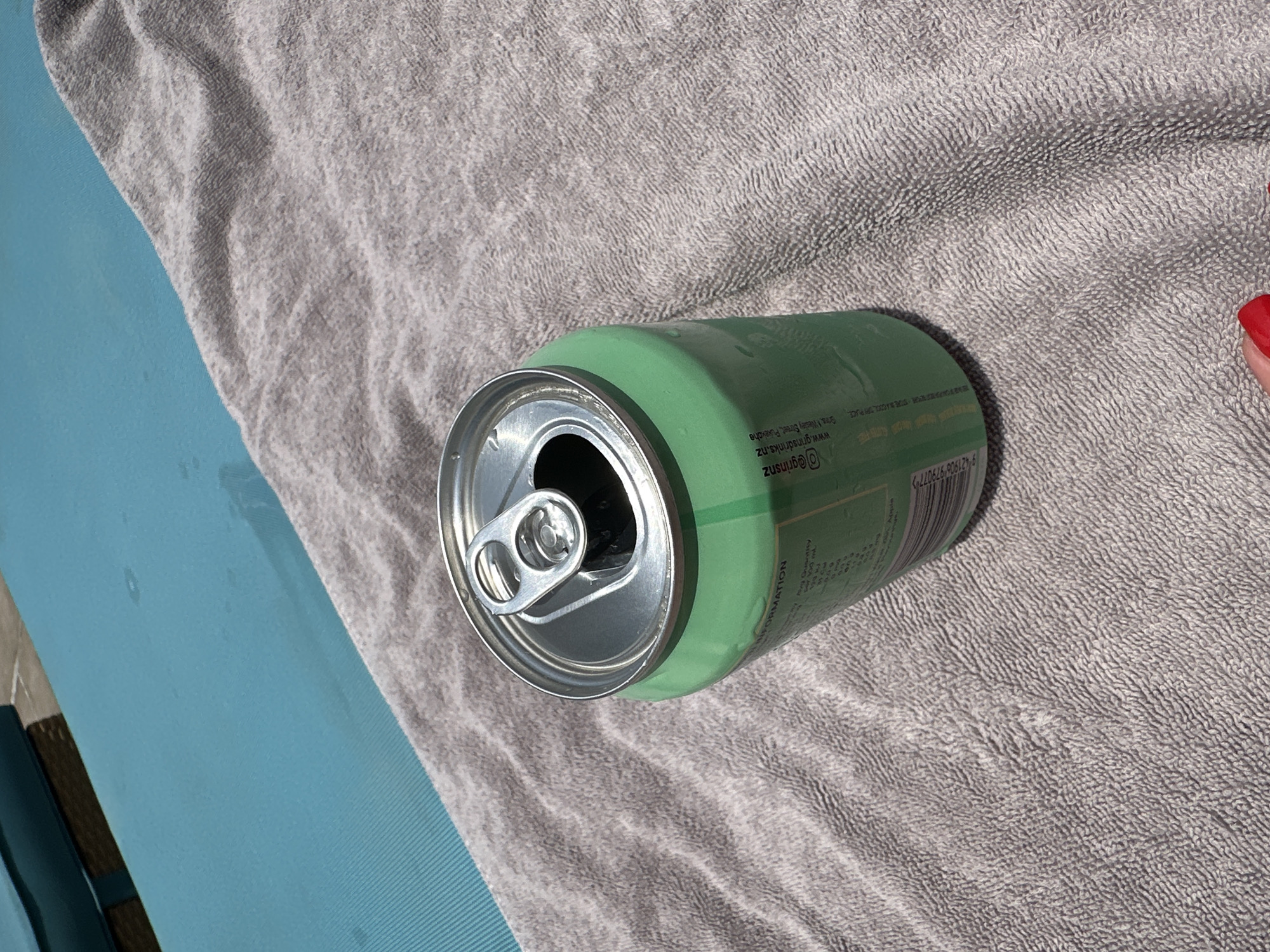 A green soda can sits open on a gray towel, which is placed on a blue chaise lounge. The chaise lounge appears to be poolside, as a faint reflection of water can be seen on its surface. The scene is brightly lit by natural sunlight. The background is blurred and out of focus. No people are visible in the image. There is no information to suggest this image was taken in Dubai, United Arab Emirates.