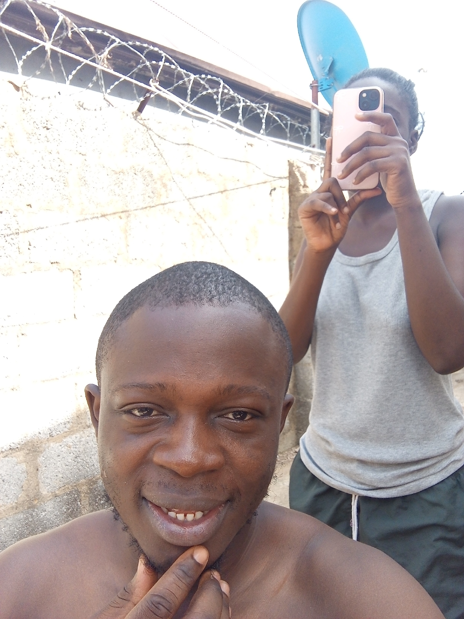 Two individuals are visible in the image. The foreground features a dark-skinned man, likely in his late 20s or early 30s, partially shirtless and with short hair. He is smiling and touching his chin with his right hand. Behind him stands another individual, also dark-skinned, holding a pink smartphone up to their face, presumably taking a photograph. They are wearing a grey tank top and dark green shorts. In the background, there is a concrete wall topped with barbed wire. A blue satellite dish is mounted above the wall. The location appears to be outdoors, possibly in a residential area. Due to the absence of distinctive landmarks, specific identification of a location within Abuja, Nigeria, is not possible.