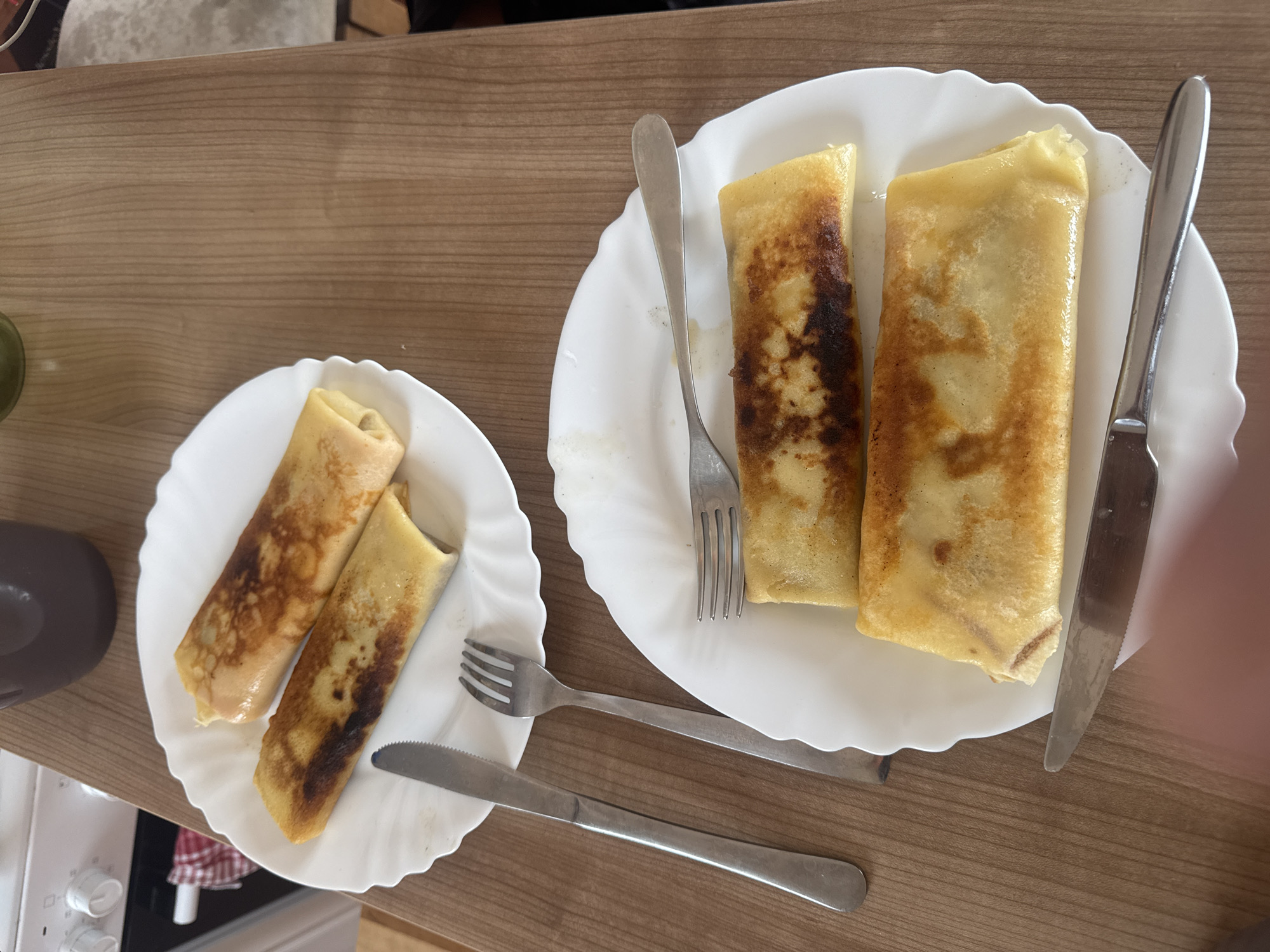 On a wooden surface are two white plates, each containing two rolled crepes, accompanied by a fork and a knife. The crepes are golden brown, with some having darker patches. The plates have a scalloped edge. The silverware is stainless steel. Part of a stove and a brown object are visible in the background. The location appears to be an indoor setting, possibly a kitchen or dining area. The city and specific location are unknown.