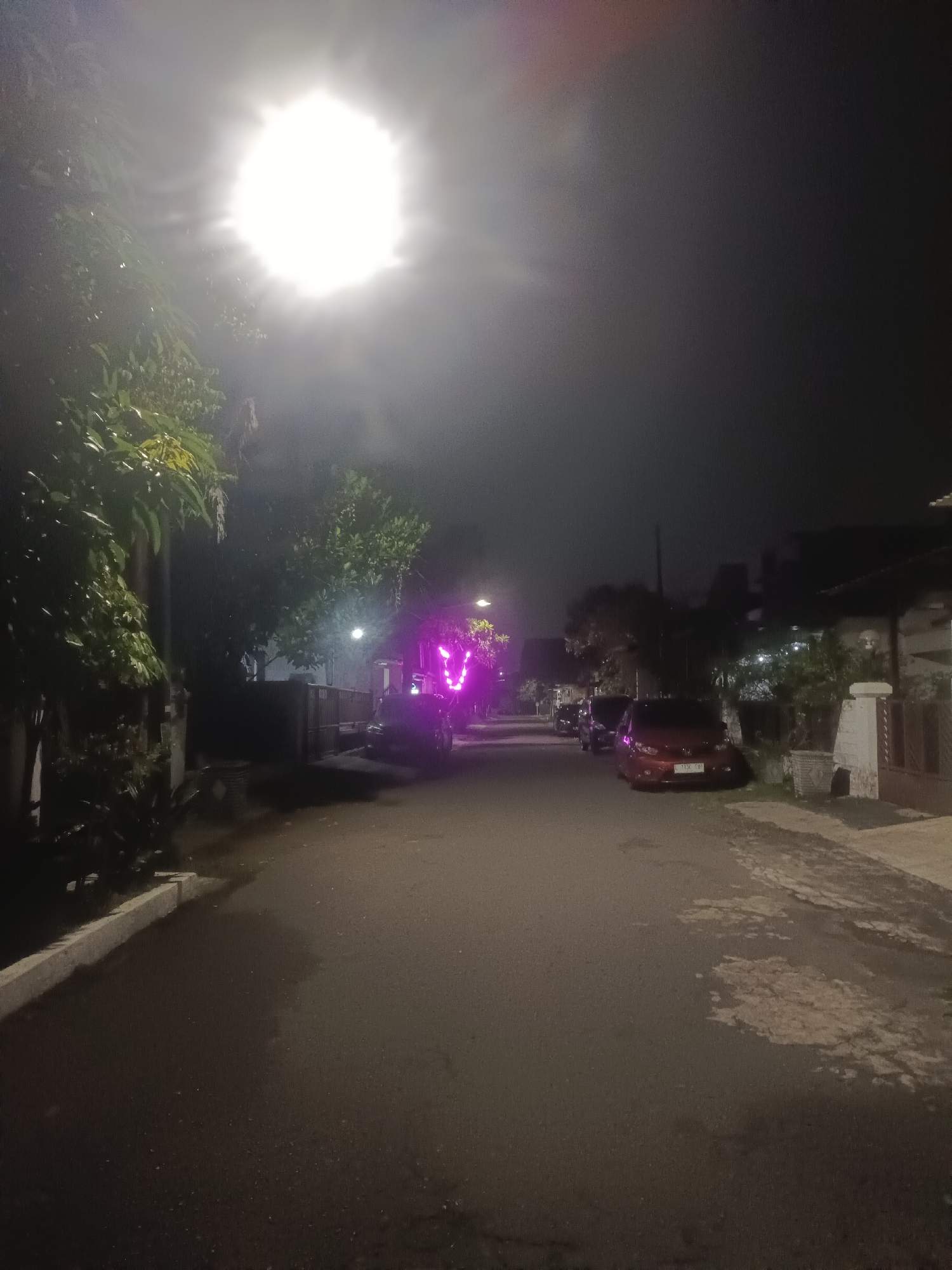 The image depicts a dimly lit residential street at night. The street is paved and stretches into the distance, flanked by trees and houses. A bright, possibly artificial, light source is prominent in the upper part of the image, obscuring details. Further down the street, there are purple lights emanating from an unknown source. Several parked cars are visible on the right side of the street. The environment suggests an urban or suburban neighborhood.