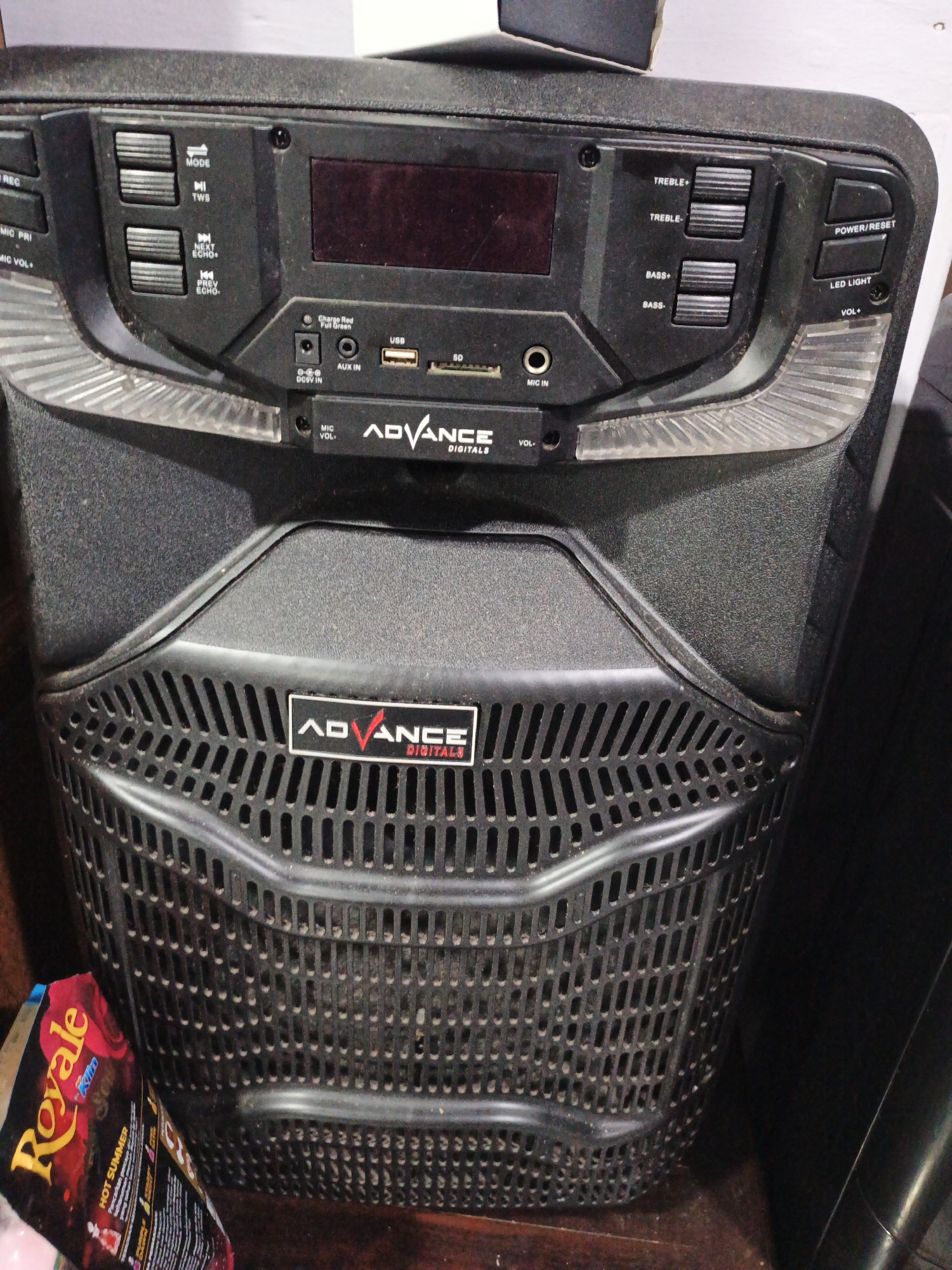 A black portable speaker with an electronic display and various control buttons is the primary subject in this medium shot, low-angle image. The speaker is positioned on a dark, wooden surface. To the left of the speaker, a corner of a brightly colored product packaging, featuring yellow text that reads "Royale" and other unreadable text, is visible. The speaker has a large grille on its lower half and a control panel with buttons for "MODE," "TWS," "NEXT ECHO," "PREV ECHO," "TREBLE+," "TREBLE-," "BASS+," "BASS-," "POWER/RESET," and "LED LIGHT." It also features ports for USB, AUX IN, DC 5V IN, and MIC IN. The "ADVANCE" logo is prominently displayed on the speaker. The background is dark and out of focus, suggesting an indoor setting. The lighting appears to be dim. There are no people or any actions occurring in the image. The location is identified as Kelapapati, Indonesia, though this specific detail is not visually verifiable from the image content.