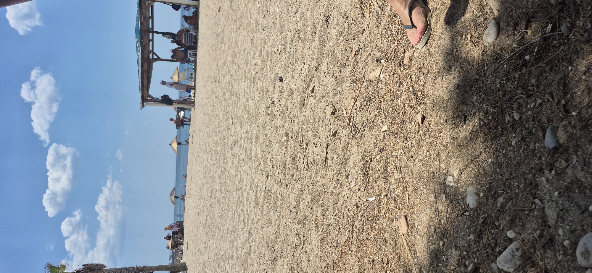 The image depicts a sandy beach scene in Paleo Faliro, Greece, under a bright blue sky with scattered white clouds. The foreground is dominated by coarse, light-brown sand littered with small pebbles, dried twigs, and other organic debris. A person's foot, wearing a flip-flop with pink toenails, is visible in the lower right corner. In the midground, several beach umbrellas provide shade for people relaxing on lounge chairs and benches. A wooden, open-sided structure, possibly a bar or cafe, offers more sheltered seating for a group of individuals. The sea is visible in the background, with a calm surface. A palm tree stands on the far left, partially framing the scene. The overall atmosphere suggests a leisurely day at the beach.