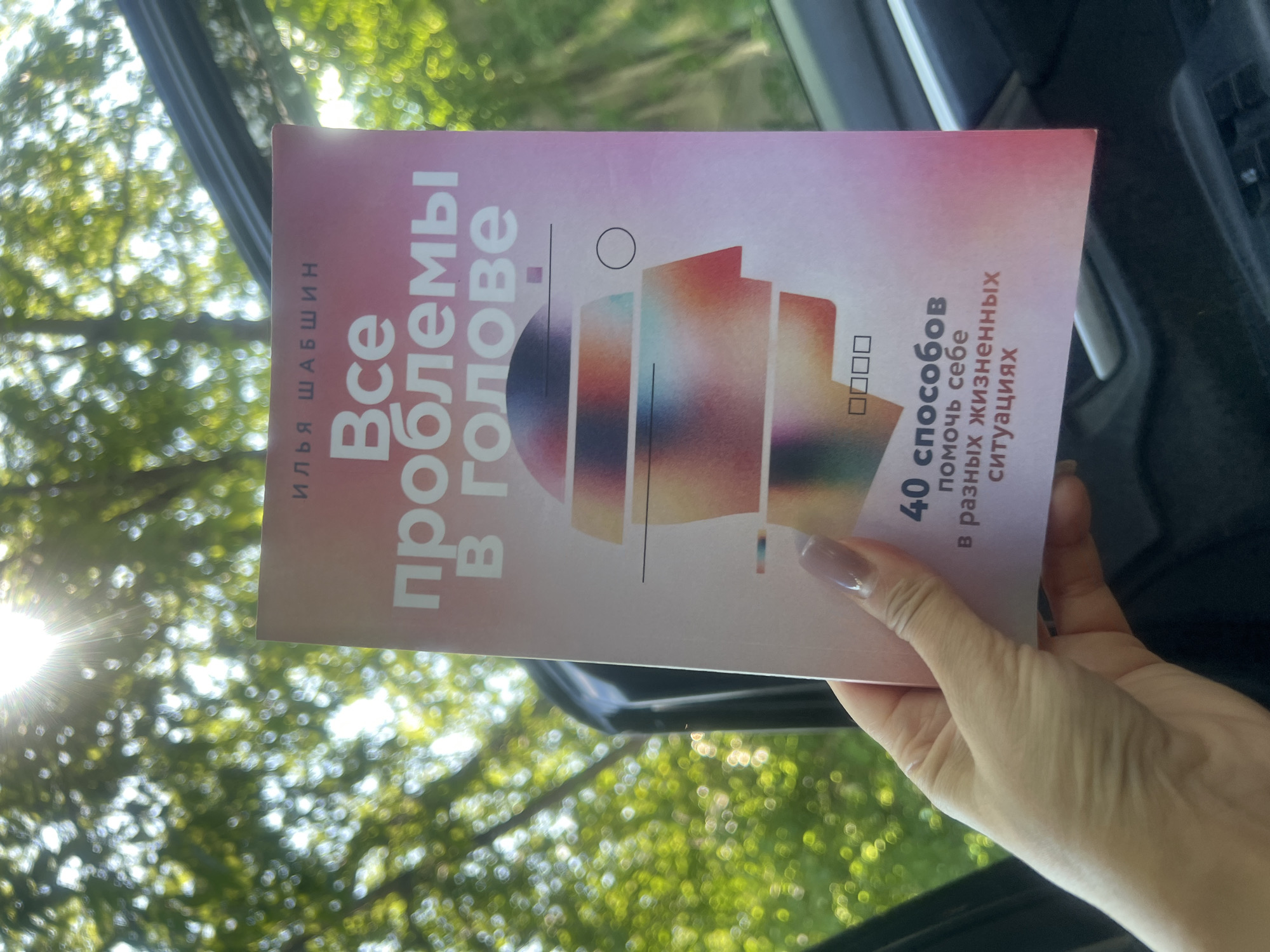 A person's hand holds a book titled "Все проблемы в голове" (All problems are in the head) with the author Ilya Shabshin. The book has a pink gradient cover with an abstract illustration of a human head with layers. Below the title, it says "40 способов помочь себе в разных жизненных ситуациях" (40 ways to help yourself in different life situations). The hand holding the book has neatly manicured nails. The background is blurred green foliage with sunlight filtering through, suggesting an outdoor setting, possibly near trees. The scene appears to be captured from inside a car, with a sunroof visible in the upper part of the image.