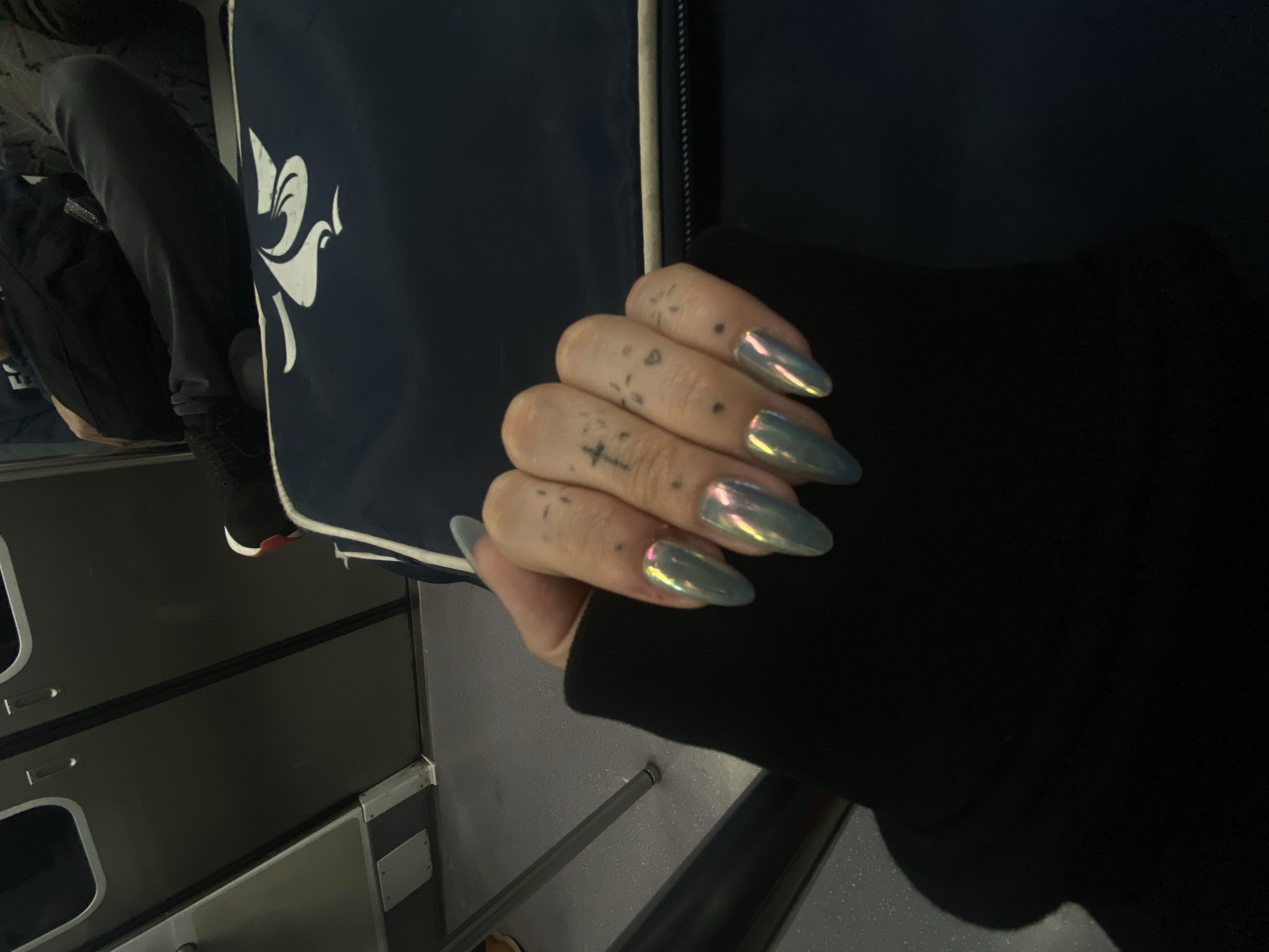 A close-up shot depicts a hand with iridescent almond-shaped nails, each finger adorned with minimalist tattoos, wearing a black long-sleeved garment. The hand rests against a navy blue surface featuring a stylized white bird design. In the blurred background, one can discern elements of an airplane interior, including seats, overhead compartments, and window frames. The setting appears to be inside an aircraft, not specific to the city of Buenos Aires, Argentina.