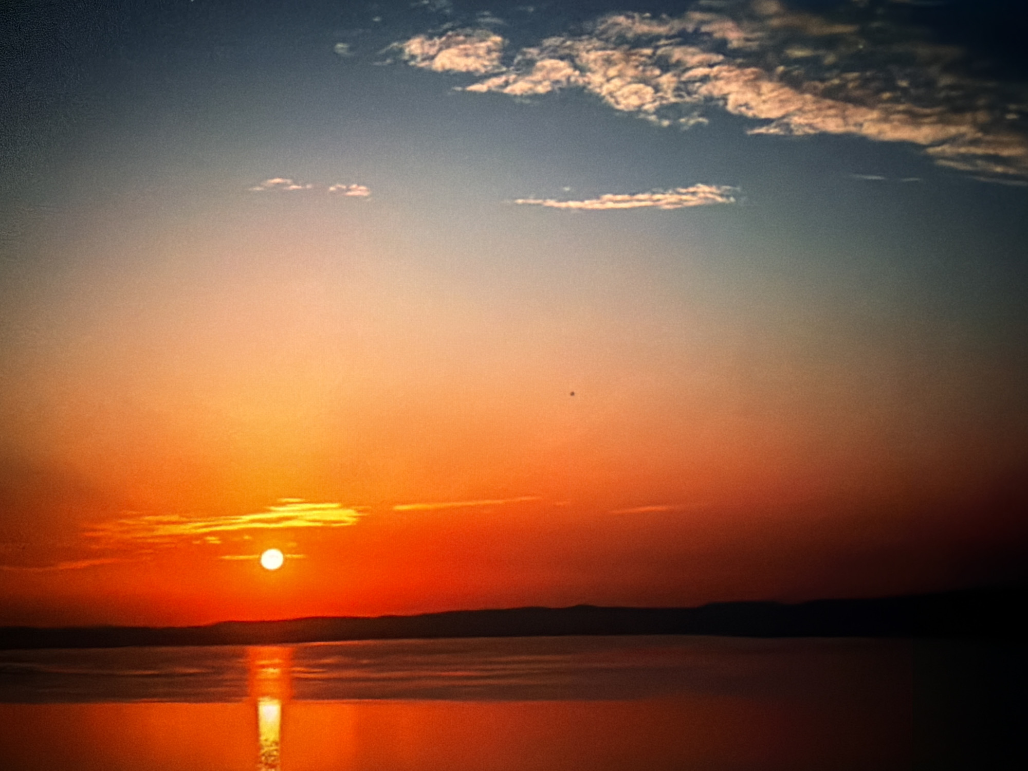 The image depicts a sunset or sunrise scene over a body of water, set in Maidstone, United Kingdom. The sky transitions from a dark blue and grey at the top, through lighter blue and peach tones, to an intense orange and red gradient near the horizon. Scattered, elongated clouds, illuminated by the warm light of the sun, are visible across the upper sky. A small, dark, circular speck is faintly discernible in the upper-mid right section of the sky.
A bright, circular sun, appearing white-yellow, is positioned just above the horizon in the lower-left quadrant of the sky. It casts an intense red and orange glow that dominates the lower half of the sky. A dark, undulating silhouette of a distant landmass or hills stretches horizontally across the horizon line, visible between the water and the vibrant sky.
The body of water below strongly reflects the sun's light, forming an elongated, vertical column of bright orange and red. The rest of the water's surface is a deep reddish-orange, gradually becoming darker towards the foreground and sides. There are no discernible people, vessels, or specific man-made objects within the frame. The scene is devoid of explicit actions or interactions.