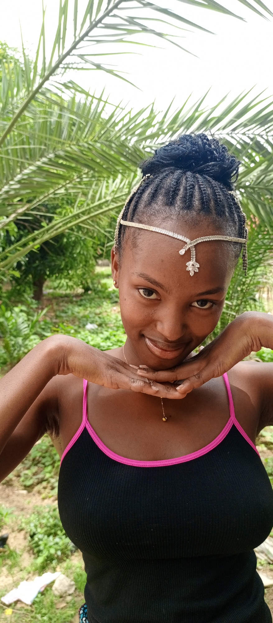 A young woman with dark skin and dark braided hair, styled in a bun, is captured in a close-up, outdoor portrait. She is wearing a black ribbed tank top with vibrant hot pink trim on the straps and neckline. A delicate, shimmering headband or headpiece, adorned with small jewels and a central leaf-shaped crystal design, rests on her forehead and across her hair. She also wears a thin gold necklace with a small, round pendant and small gold stud earrings. Her fingernails have a light-colored polish.
The woman is posing for the camera, smiling gently with a direct and engaging gaze. Her hands are clasped together, and her chin rests upon them, creating a sweet and slightly coy expression.
The background consists of lush green foliage, with large palm-like leaves prominently featured, suggesting a tropical or subtropical environment. Other green trees and plants are visible further back. The ground beneath the foliage appears to be unpaved earth with scattered green growth. The bright, natural lighting indicates it is daytime. No text is visible in the image.