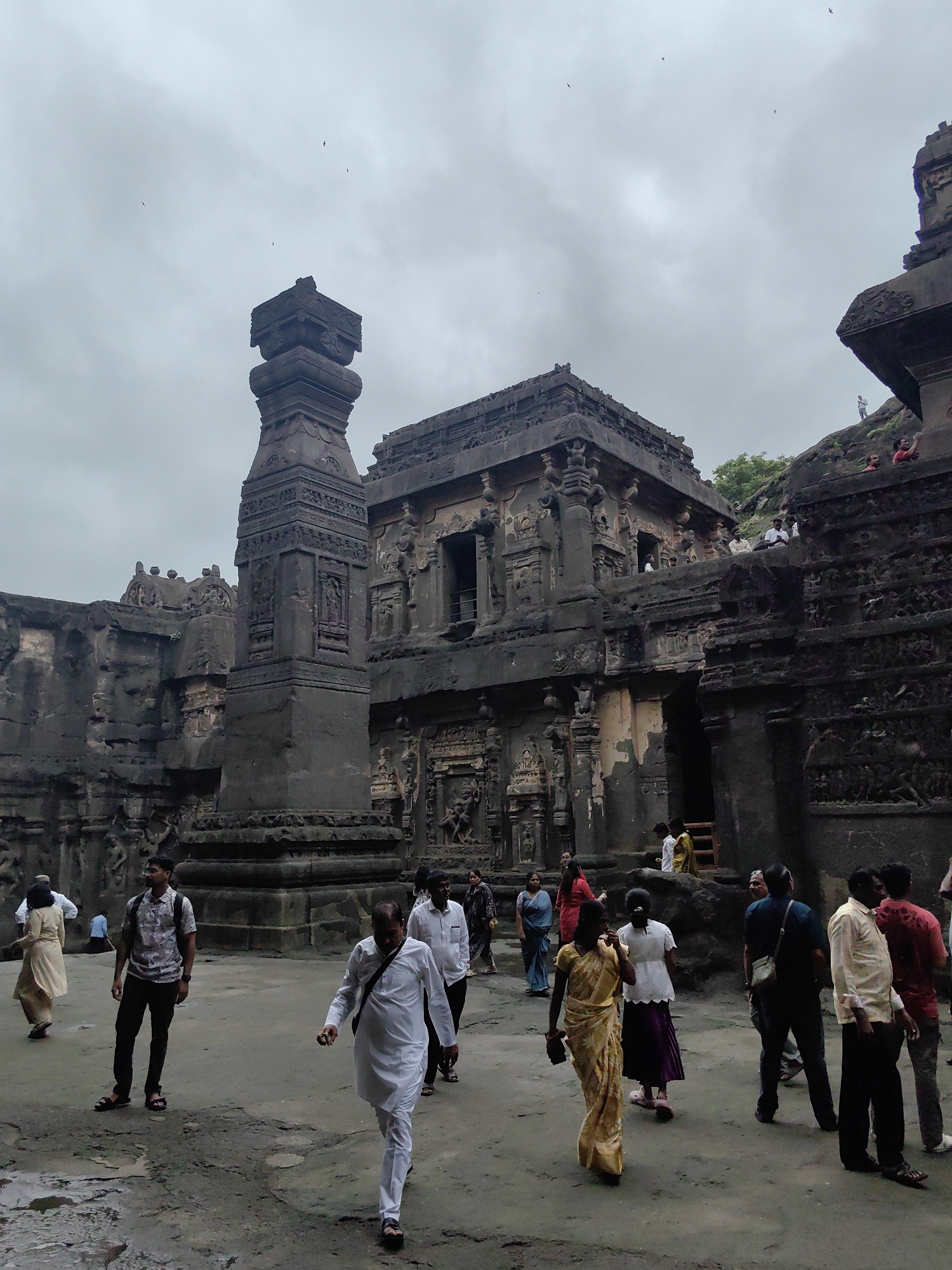In Ellora, India, a group of tourists are exploring a vast, ancient rock-cut temple complex under a cloudy sky. The massive structures, carved directly from the cliff face, feature intricate sculptures and architectural details, showcasing a significant historical and cultural site. Several individuals are walking on the uneven, stony ground, some observing the monumental architecture, while others are captured in candid moments of movement. The scene depicts a lively and engaging tourist experience at this remarkable UNESCO World Heritage site.
