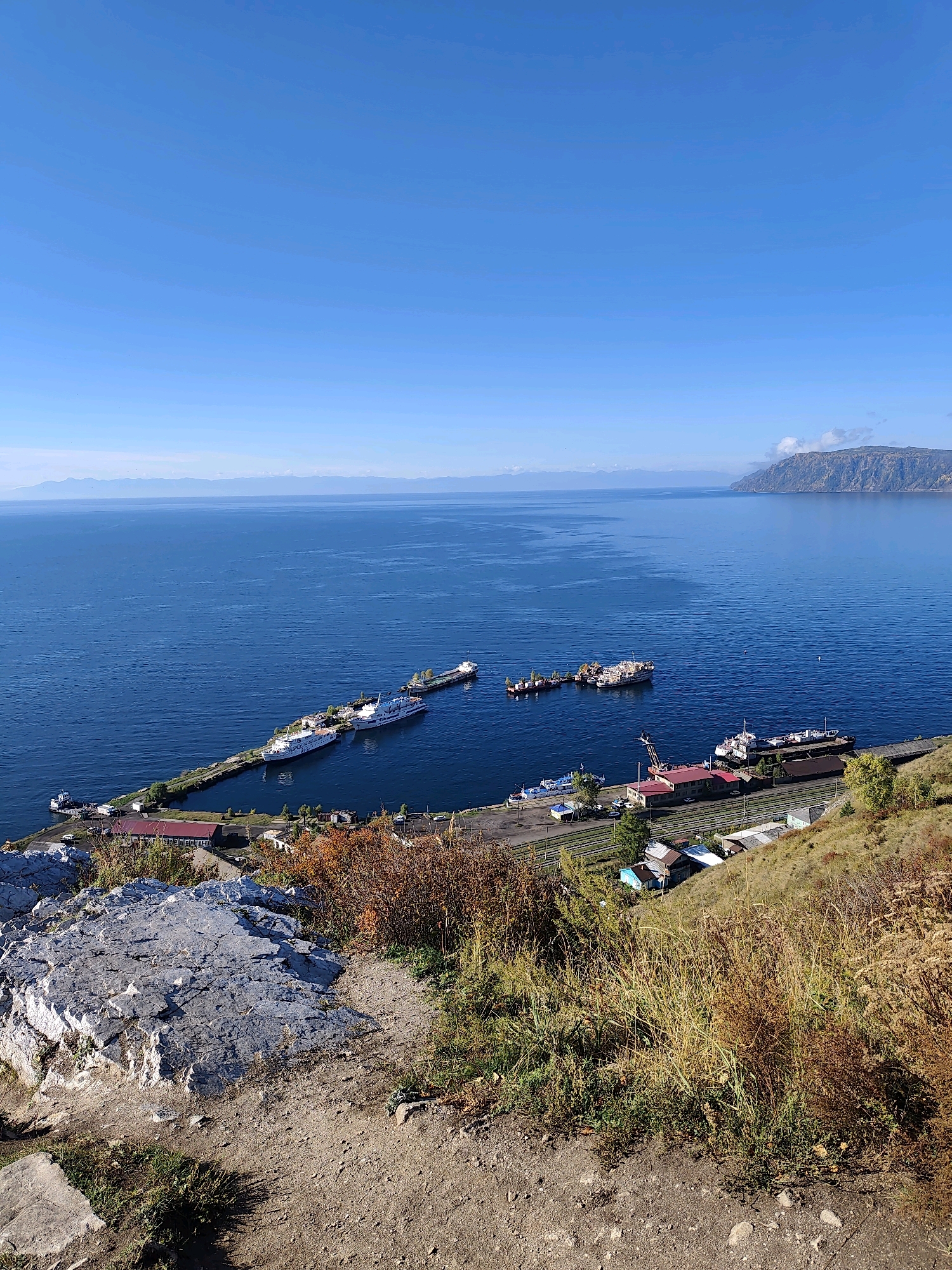 This is a scenic, elevated, eye-level, wide shot of a calm, deep blue body of water, likely Lake Baikal in Russia, under a clear, bright blue sky. In the mid-ground, a long pier juts out from the shore, with several boats docked alongside it, including a large white passenger ferry and a smaller tugboat. Along the shore, there is a small settlement with various buildings, some with red roofs, and a train track running parallel to the water. The foreground is dominated by a rocky, dry, and grassy hillside, with sparse autumnal foliage. In the distance, the water stretches to the horizon, meeting a range of hazy, mountainous landforms. The scene captures a peaceful, somewhat remote, coastal or lakeside environment.