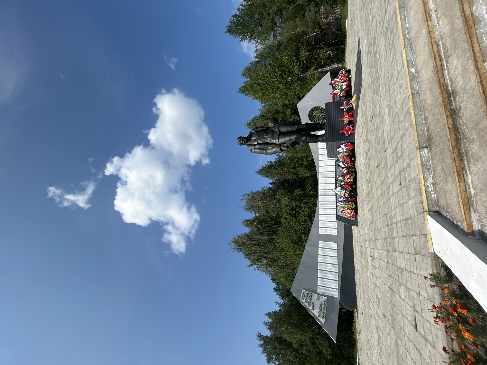 The image shows a war memorial in Krasnovishersk, Russia, under a bright blue sky with scattered white clouds. The memorial features a large, dark statue of a man in uniform, holding something at his side. The statue stands on a dark base in front of a gray, angular monument. This monument has rows of white plaques and inscriptions in Russian. In front of the statue and monument, there are several wreaths laid on the ground. The memorial is situated on a paved plaza made of square gray tiles. To the left of the plaza, there is a flower bed with bright orange flowers. In the background, a dense forest of green trees forms a backdrop. The scene appears to be a solemn commemoration site.