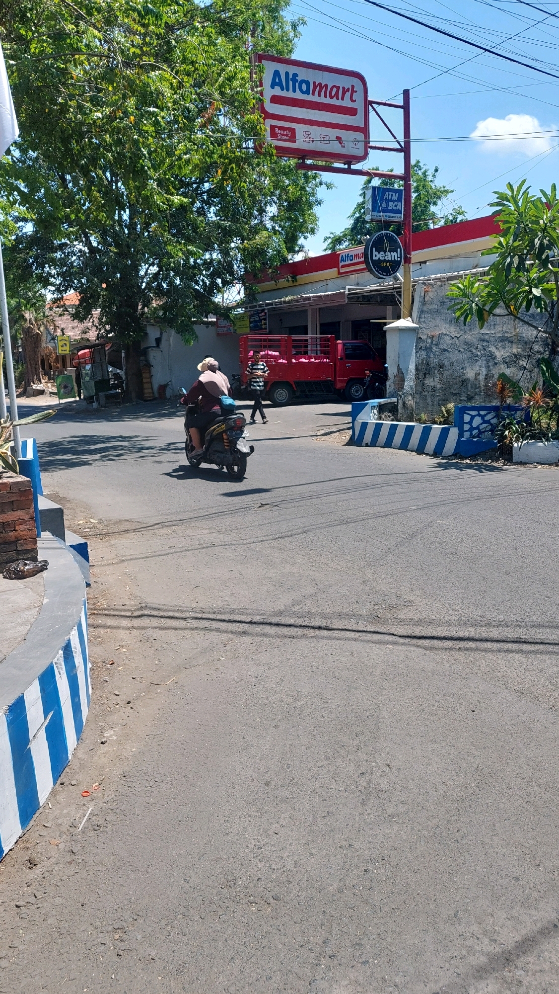 The image depicts a sunny day in Siwalan Panji, Indonesia, near an Alfamart convenience store. In the foreground, a paved road curves to the left, bordered by a concrete barrier painted with alternating blue and white stripes. A person on a scooter, wearing a light-colored hat and a dark shirt, is traveling away from the camera. Behind the scooter, a red truck with a metal bed is parked. To the right of the truck, a person in a striped shirt walks away from the store. The Alfamart store has a red and white awning, and a prominent red and white sign with the Alfamart logo is visible overhead, along with smaller signs indicating ATM and "bean!" which likely refers to a coffee or snack service. Trees with green foliage are present, and the sky is blue with some clouds. The overall scene suggests a typical street view in a commercial area of the town.